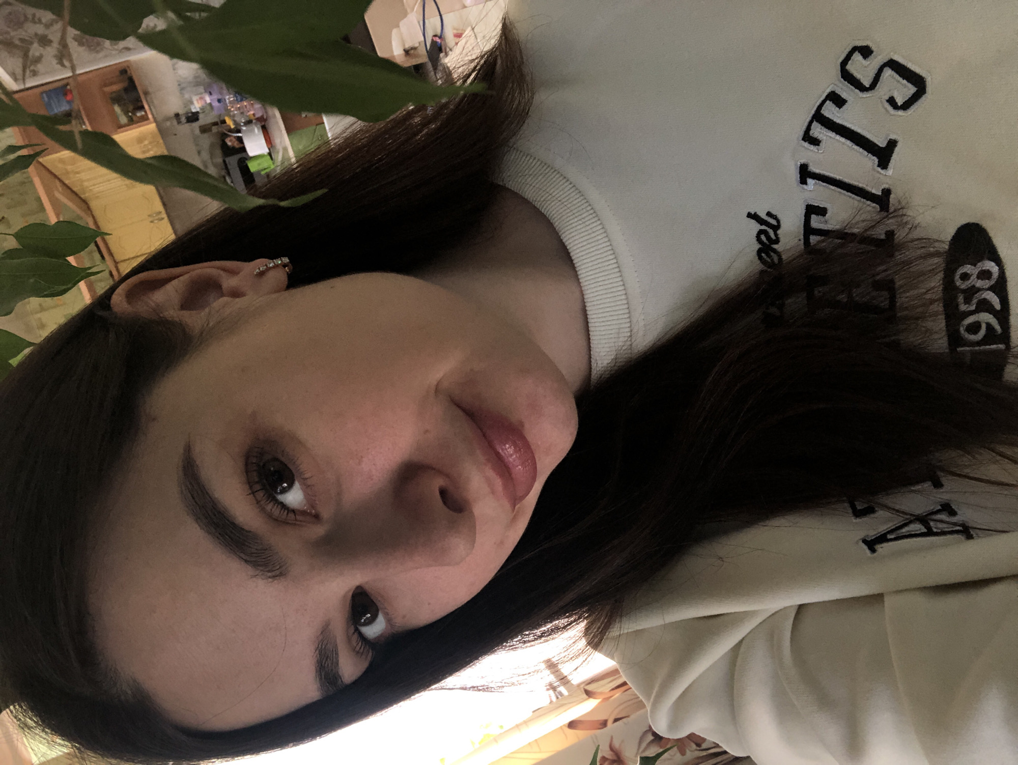 A close-up, angled selfie shows a fair-skinned young woman with dark brown hair and eyebrows. She is wearing a light-colored sweatshirt with dark lettering that reads "Sweet Titts est 1958." An earring with small, possibly crystalline, details is visible in her left ear. A leafy green plant is partially visible in the top left of the frame, slightly obscuring the background. The background includes glimpses of a domestic interior, possibly with shelves or cupboards. The location, Noviy, Russia, cannot be confirmed from this image.