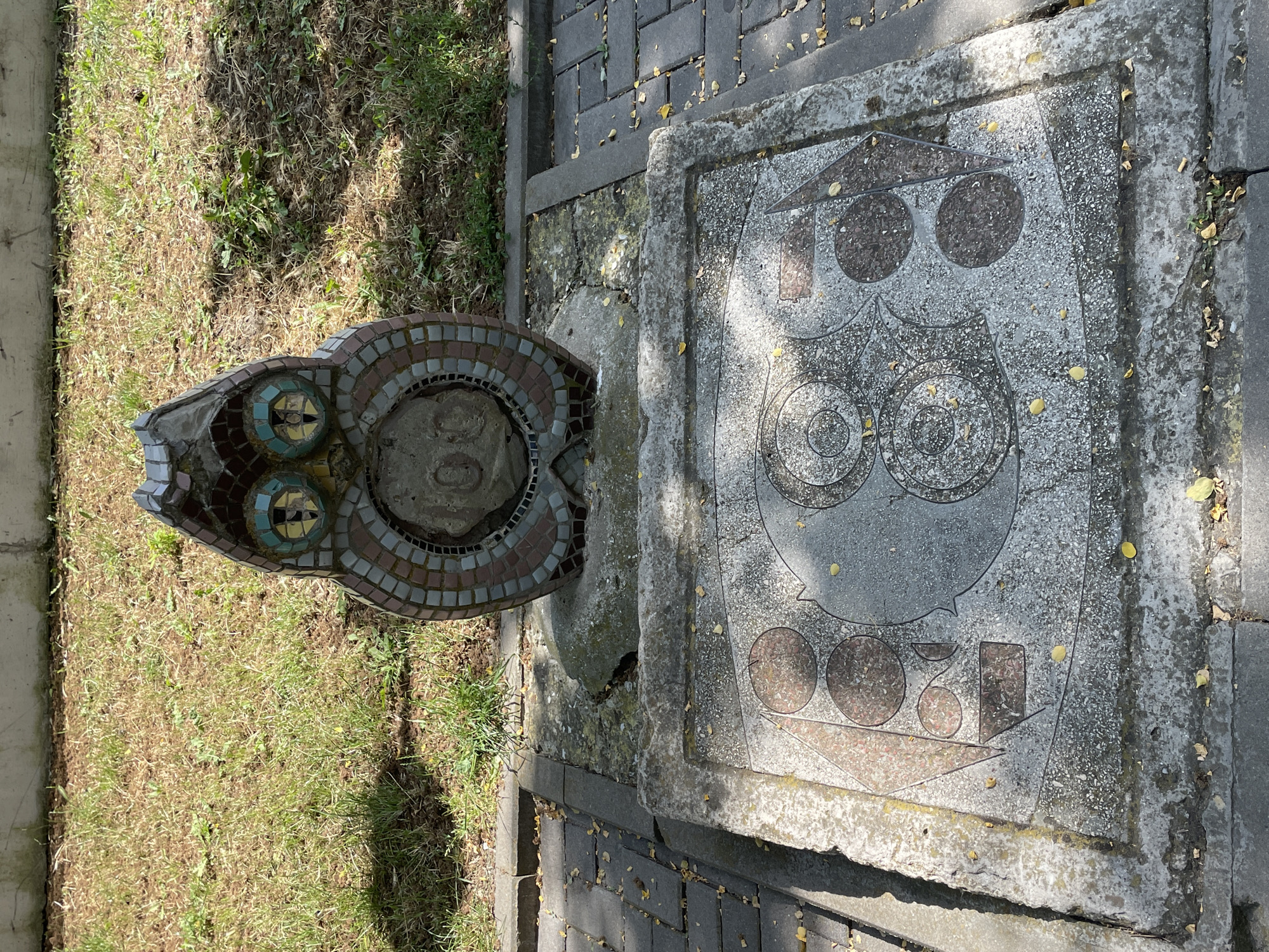 This media file depicts an outdoor scene featuring a mosaic sculpture and a stone carving. The sculpture, shaped like an animal's head with colorful mosaic tiles forming its eyes and body, is positioned to the left of the frame. To the right, a large, rough-hewn stone slab is carved with a stylized owl motif. The owl design includes large, concentric circles for eyes and a triangular head, all set within a stone frame.
The ground surrounding the sculpture and carving is a mix of short, dry grass and a paved walkway made of dark grey interlocking bricks. Scattered yellow leaves are visible on the grass and the paving stones, suggesting it might be autumn. The lighting suggests it is daytime, with shadows cast by the objects. The scene appears to be in a public outdoor space, possibly a park or garden, given the presence of both grass and paved pathways. There is no visible text in the image. The overall impression is one of a quiet, perhaps slightly weathered, outdoor art installation.