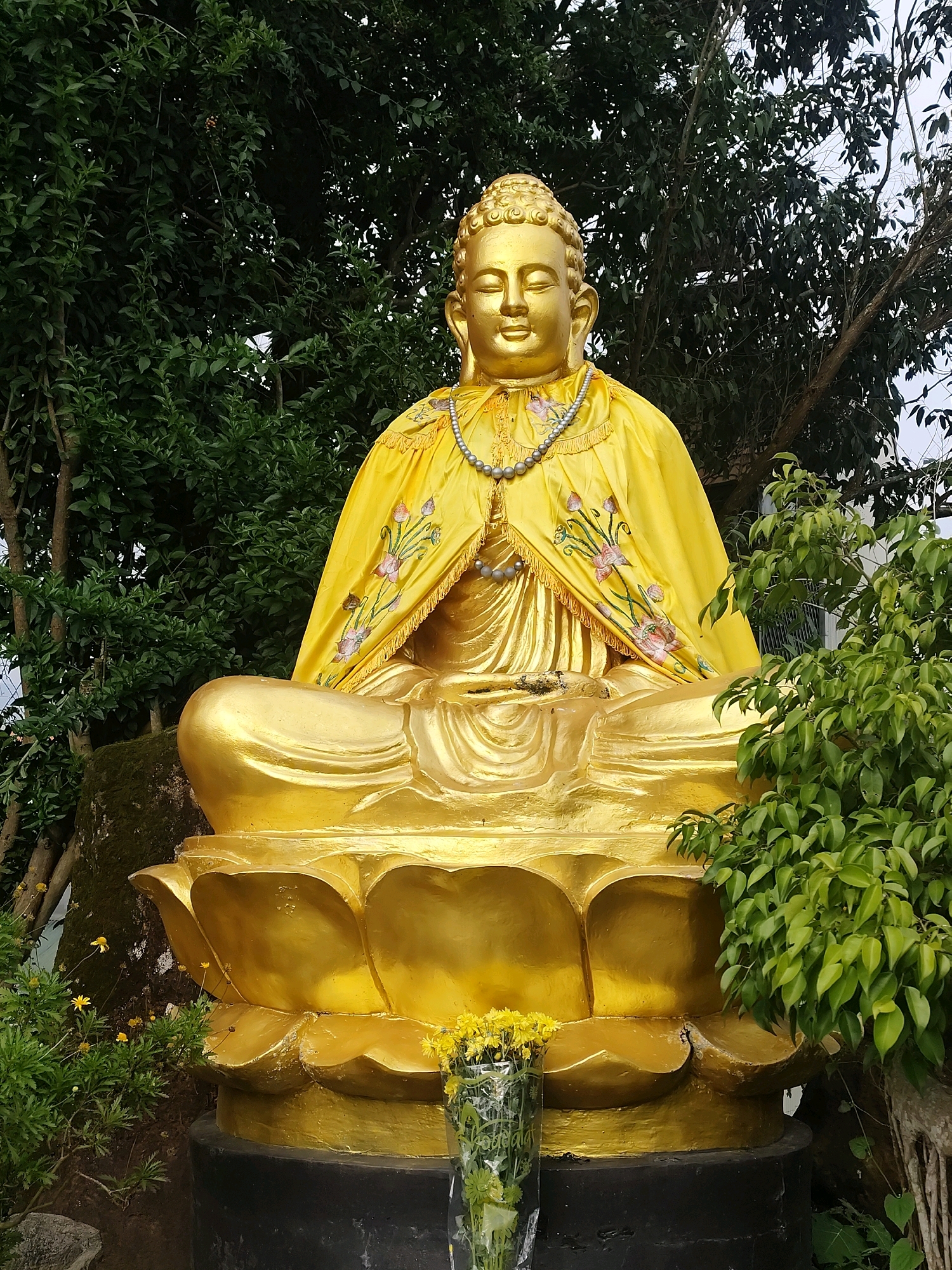 A large, golden statue of Buddha sits in a meditative pose on a lotus flower pedestal. The Buddha is adorned with a yellow robe embroidered with pink and white flowers, and a pearl necklace. Green trees and foliage surround the statue, with a glimpse of a grey sky through the leaves. A clear vase filled with yellow flowers is placed in front of the lotus pedestal. The setting appears to be an outdoor religious or cultural site in Lâm Viên - Đà Lạt, Vietnam.