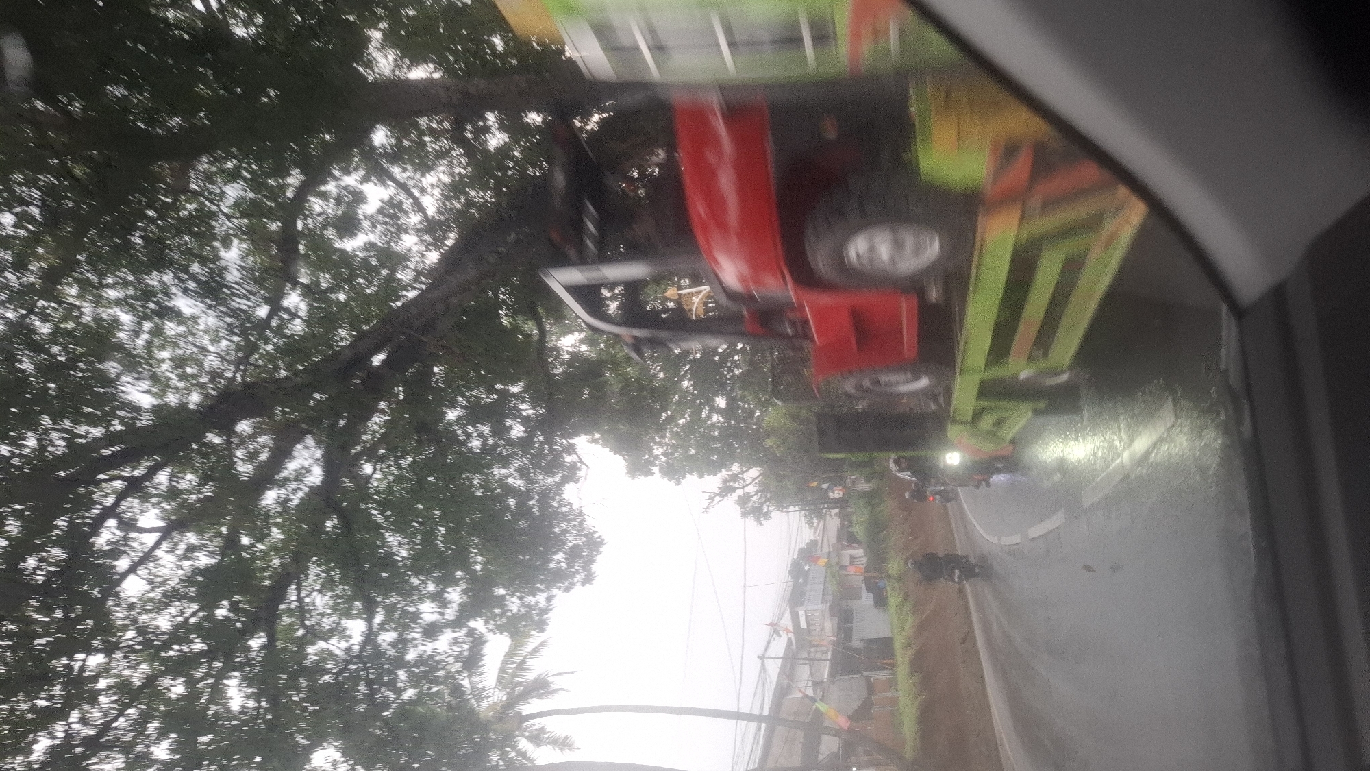 The image shows a street scene in Cilaku, Indonesia, on a cloudy day, possibly after rain. A red forklift is being transported on a flatbed truck, which is on the right side of the frame, partially obscuring the view. The truck has a green bed with orange reflective markings. A large tree with dense foliage hangs over the road from the left. In the background, there are houses and palm trees. Several motorcycles are on the wet road, with one rider in the foreground on the left and another further back on the right. The overall impression is one of movement and a somewhat hazy atmosphere.