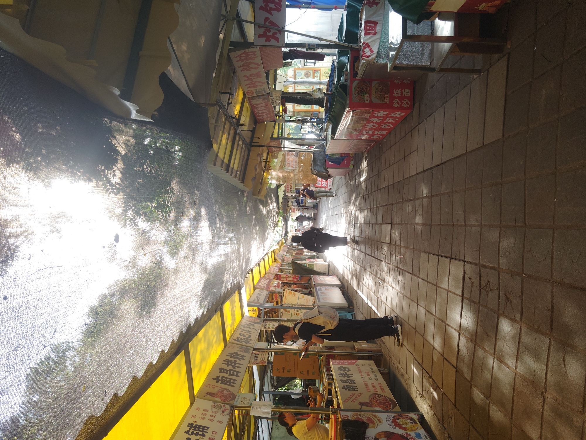 Here is a matter-of-fact description of the scene in the image:
The image shows an outdoor food vendor area, likely in Chongqing High-tech Zone, China. The stalls line a paved walkway, with awnings or covers providing shade. The signs are in Chinese, advertising various food items, including noodles, fried rice, and desserts like "Bingfen" (ice jelly). People are present, including a vendor preparing food and customers browsing the stalls. Some people are walking down the path. The light is bright, creating strong shadows.