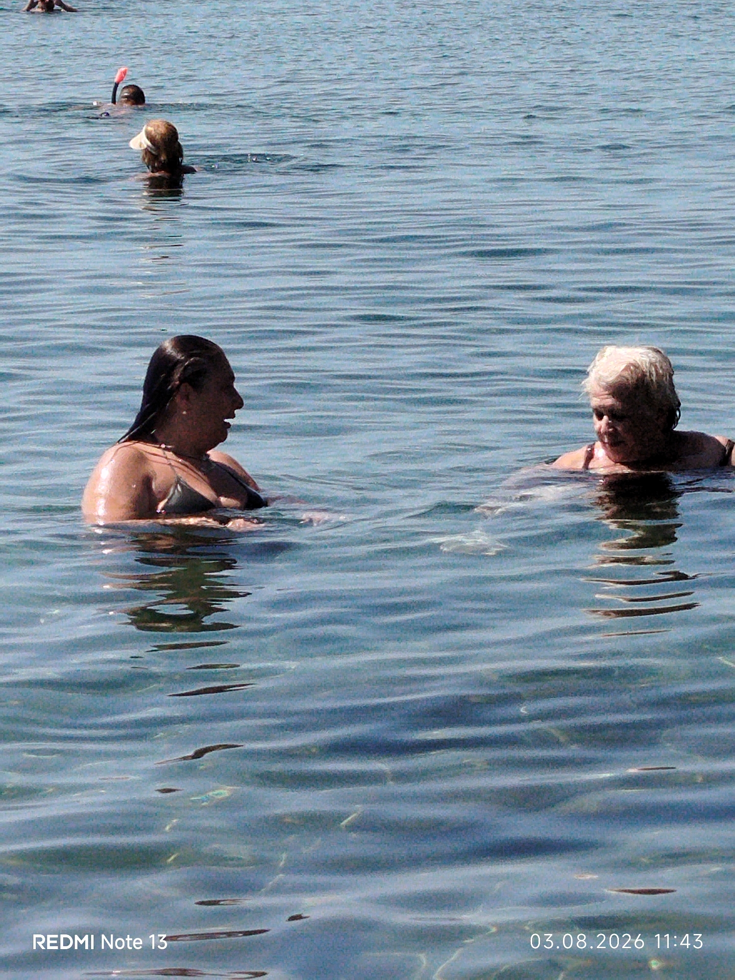 Two women are swimming in the sea in Sithonia Municipal Unit, Greece. The woman on the left is facing right, with her mouth open and her body partially submerged. She is wearing a dark bikini top. The woman on the right is facing left, with her head tilted down. She has short, blonde hair and is wearing a light-colored swimsuit. The water is clear and blue, with some ripples on the surface. Other people are visible in the background, some swimming and some with snorkels. The bottom right corner of the image displays the text "03.08.2026 11:43" and "REDMI Note 13".