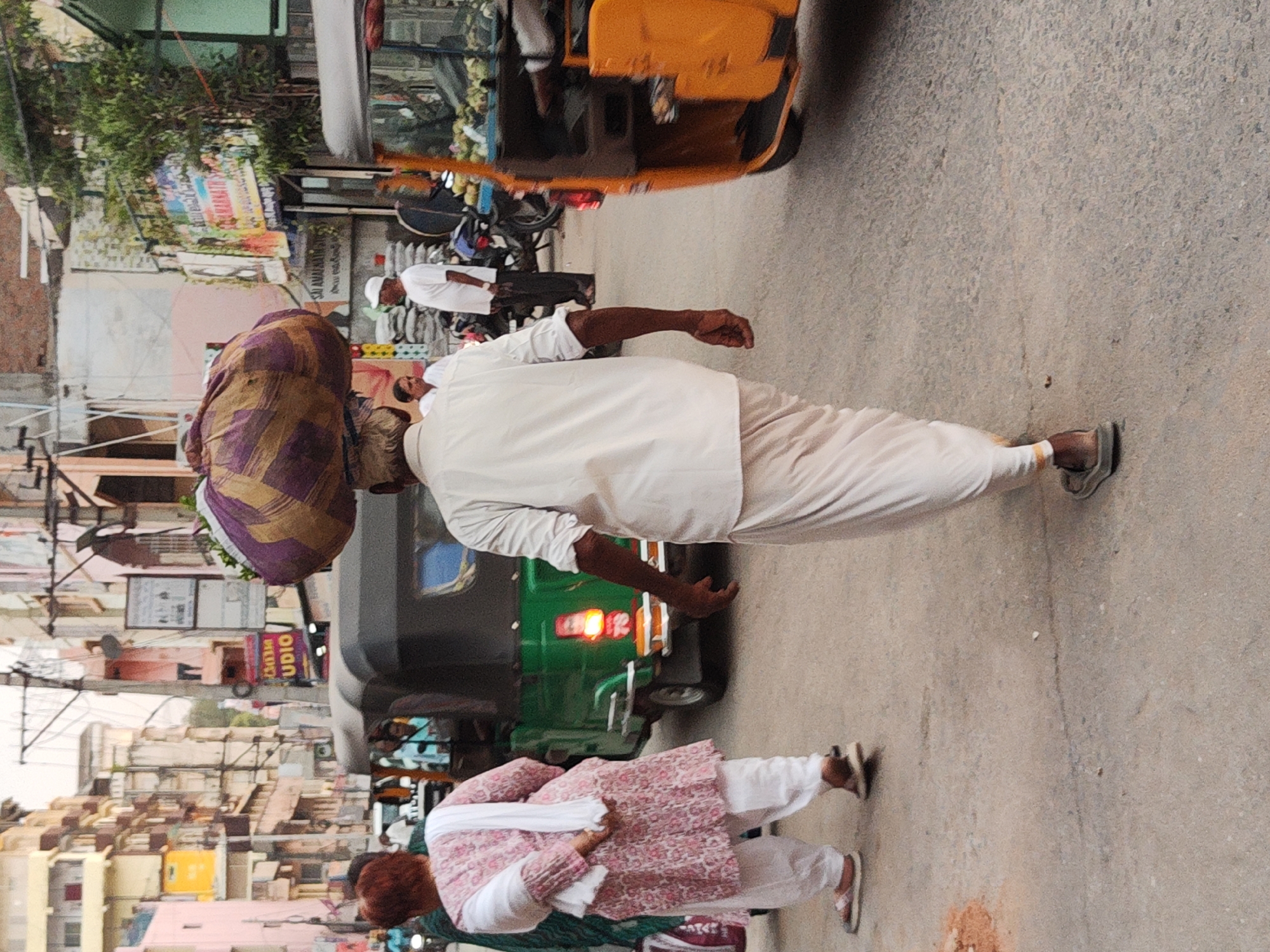 In Puttaparthi, India, a man in white attire carries a large, striped bundle on his head, walking along a street adjacent to auto-rickshaws. Another man, also in white, stands nearby. A woman in a pink and white patterned outfit walks in the same direction. The scene shows a typical urban street setting in India.