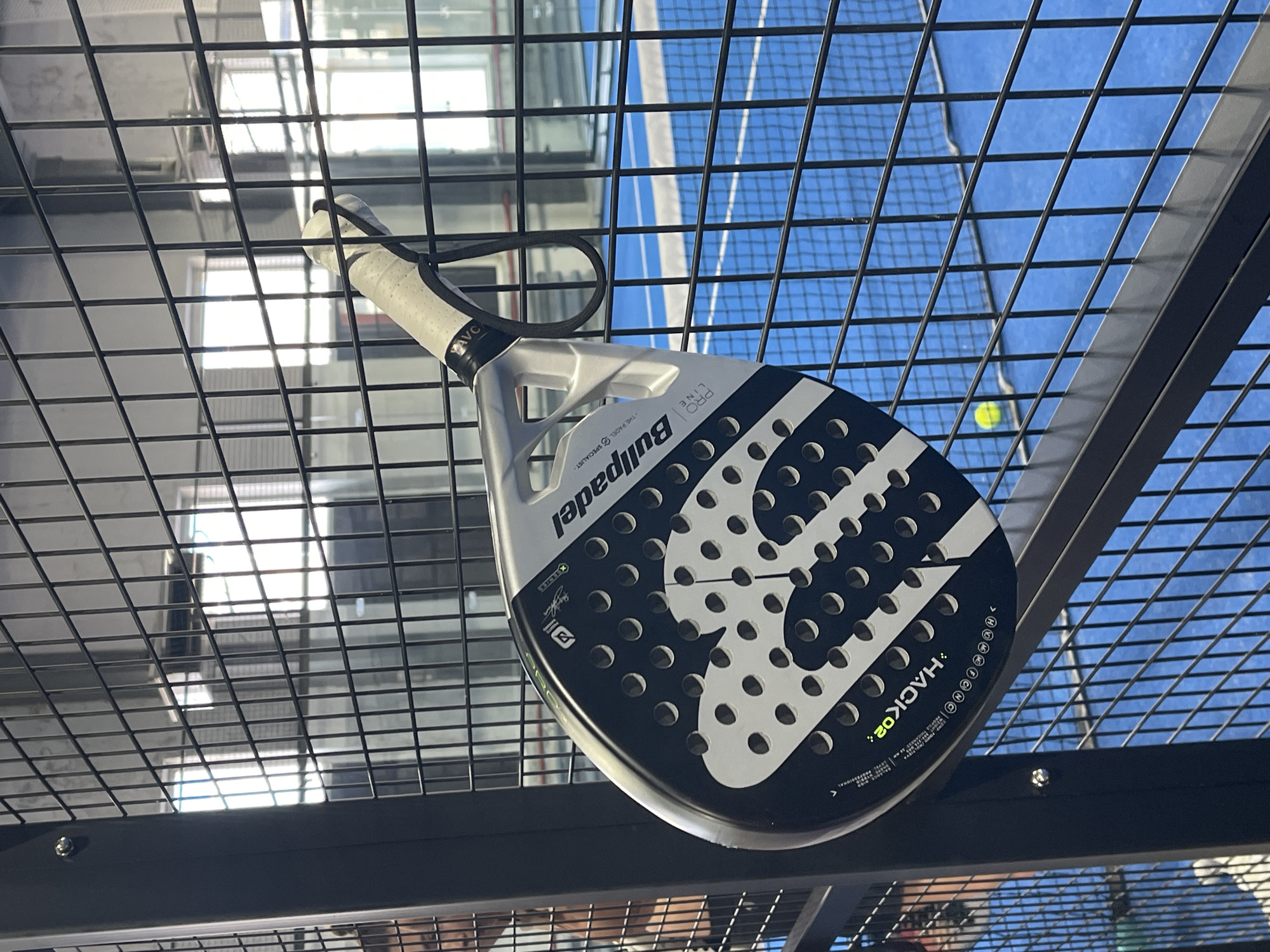 The image shows a black and white padel racket hanging on a metal fence. The racket is positioned diagonally, with the handle at the top right and the head at the bottom left. The brand "Bullpadel" is visible on the racket head, along with the model name "HACK 02". In the background, a blue padel court with a net is visible, along with a yellow padel ball. The setting appears to be an indoor sports facility. There are no identifiable landmarks to suggest the location is Orenburg, Russia.