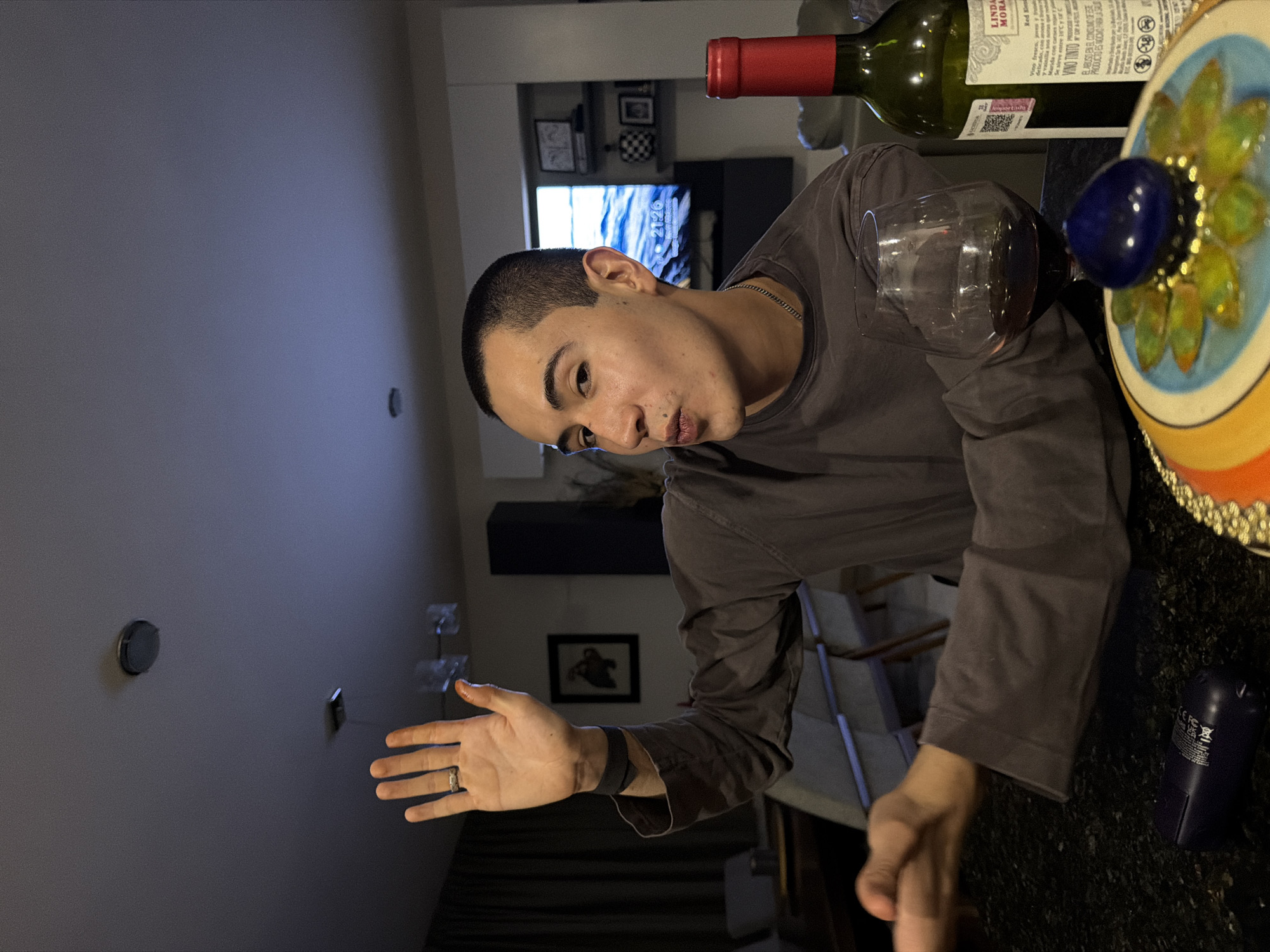 A man is in a room with wine, a wine glass, and decorative objects on a table. The time on a television in the background reads 21:26. The location is indicated by the wine bottle label as Mexico.