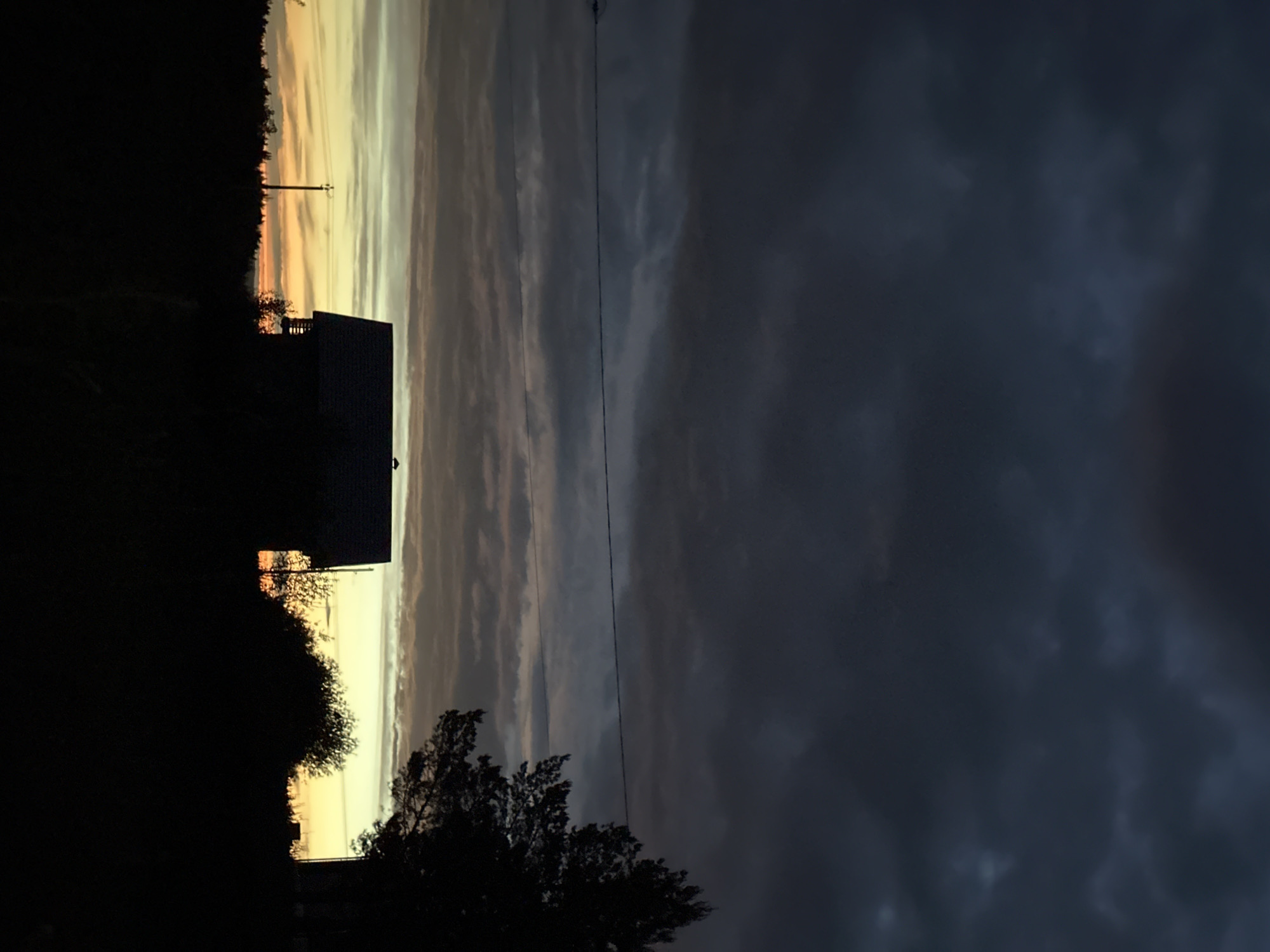 The image depicts a dusk or dawn scene in Прикамский, Russia. A dark silhouette of a house with an adjacent tree occupies the left side of the frame. Behind the house is a band of golden light on the horizon, likely indicating the sun's position. Further to the right, the sky transitions into darker, cloudy formations. A power line stretches across the sky above the house and trees. The environment appears to be a residential area, with a dense, dark background on the left suggesting a forest or wooded area.