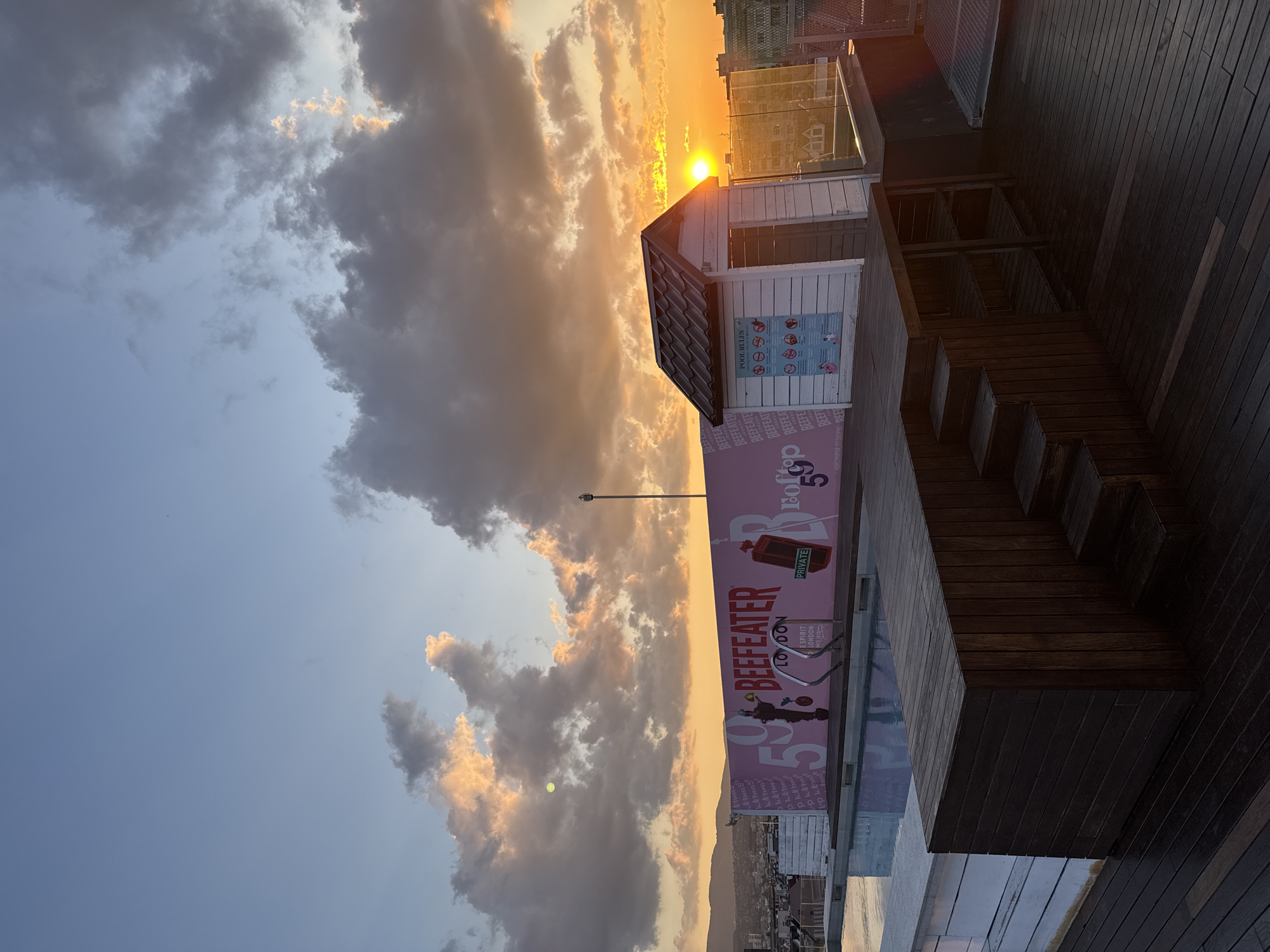 This image captures a panoramic view of a rooftop bar and pool area at sunset, overlooking the city of Sofia, Bulgaria. The sky exhibits a gradient of colors, transitioning from bright orange and yellow on the horizon, where the sun is visible as an intense disc on the far right, through soft pinks and purples, to lighter blue overhead. Scattered cumulus clouds are illuminated from below, displaying warm golden and orange hues.
In the mid-ground, a rectangular swimming pool with light blue water and a white-tiled edge is positioned on the left. Behind the pool, a prominent pink wall acts as a backdrop, featuring bold white and red branding that reads "BEEFEATER LONDON." The number "59" is prominently displayed on this wall, along with smaller text such as "PRIVATE" and "SINCE 1820," and a stylized graphic of the Beefeater mascot. Two metal pool ladders extend into the water from the wall.
To the right of the pool, a small, white wooden shed with a dark brown tiled gabled roof stands. A "POOL RULES" sign, featuring multiple pictograms, is affixed to its front facade. The entire floor of the rooftop is covered in dark brown wooden decking. A raised section of this decking, with three steps leading up to it on the right, also incorporates open shelving underneath. A transparent glass balustrade lines the right edge of the rooftop, offering an unobstructed view of the urban environment.
In the background, the cityscape of Sofia stretches out, characterized by various buildings and structures, with a range of hills or mountains visible on the distant horizon to the left. No individuals are present in the scene.
Based on the distinctive "BEEFEATER LONDON" branding, the number "59," and the rooftop pool with city views, the location is identified as The Top Rooftop Bar & Pool, situated at the Sense Hotel Sofia, located in the central part of Sofia, Bulgaria.
