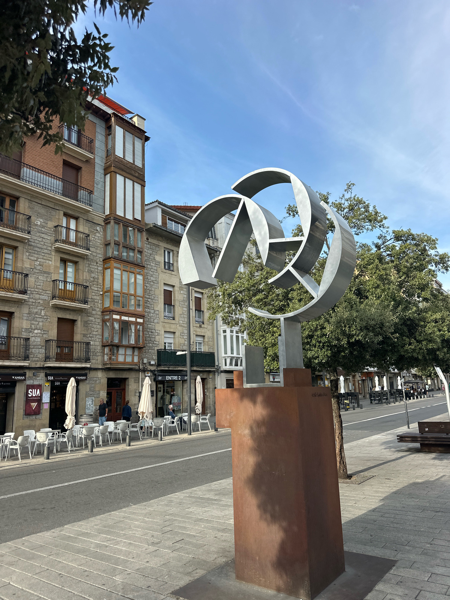 An outdoor, eye-level, wide shot captures a modern sculpture and a street scene in Vitoria-Gasteiz, Spain. The sculpture, made of weathered steel, is a series of interlocking curved metallic elements supported by a rust-colored cylindrical base. It is positioned on a sidewalk in the foreground, casting a shadow to its right.
Behind the sculpture, a two-lane asphalt street runs horizontally across the image, bordered by a paved sidewalk on both sides. The left side of the street features a row of multi-story buildings with stone and brick facades. The buildings have balconies with wrought-iron railings, large bay windows, and storefronts at street level. Umbrellas are deployed over outdoor seating areas in front of some establishments. A sign for "SUA FOOD & WINES" is visible on one of the storefronts. A few people are visible walking on the sidewalk and sitting at the outdoor tables.
The right side of the street shows a sidewalk lined with trees and more outdoor seating areas with umbrellas. A park bench is visible in the distance. The sky above is a clear blue with faint wispy clouds. The overall impression is of an urban environment with a blend of traditional architecture and contemporary art, on a clear day.