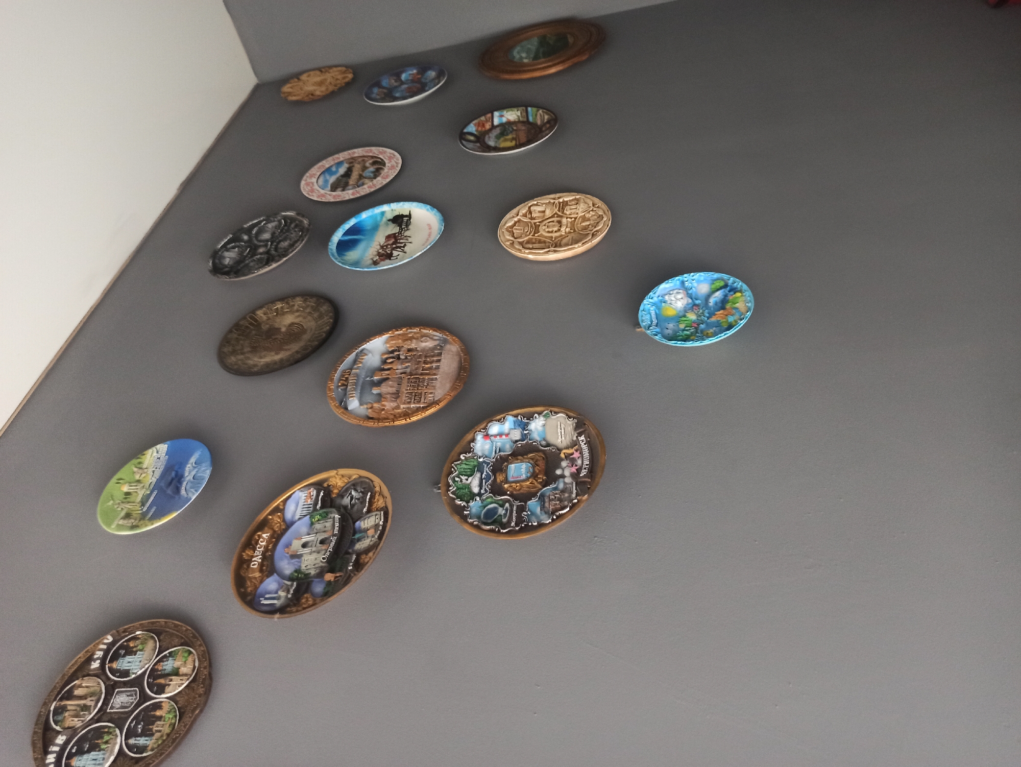 The image shows a collection of decorative plates mounted on a gray wall. The plates are of various shapes and sizes, and each appears to depict a different scene or design. Some plates are oval-shaped, while others are round or rectangular. The designs range from architectural landmarks to landscapes and abstract patterns. There are no people visible in the image. The environment appears to be an indoor setting, possibly a home or a gallery, with the plates arranged on a wall in a decorative manner. The lighting suggests it is daytime. The specific location in Kryvyi Rih, Ukraine, cannot be determined from the visual information provided.
