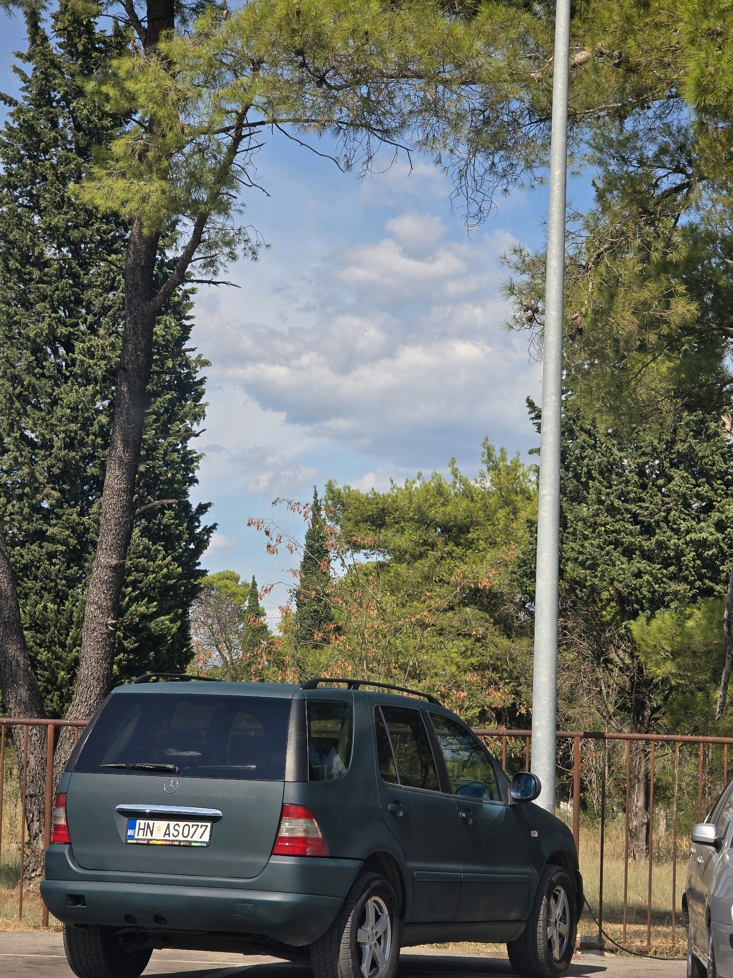 The image shows a dark green Mercedes-Benz ML-Class SUV parked outdoors, partially obscured by a metallic pole and trees. The vehicle is facing towards the viewer's right, with its rear to the left. The license plate is visible and reads "HN AS077". The background consists of lush green trees, a brown metal fence, and a blue sky with scattered white clouds. A second vehicle, possibly silver, is partially visible on the far right of the frame. The scene appears to be a parking area surrounded by vegetation, suggesting a location with a temperate climate. The specific location in Podgorica, Montenegro cannot be determined from the image alone, as there are no identifying landmarks.