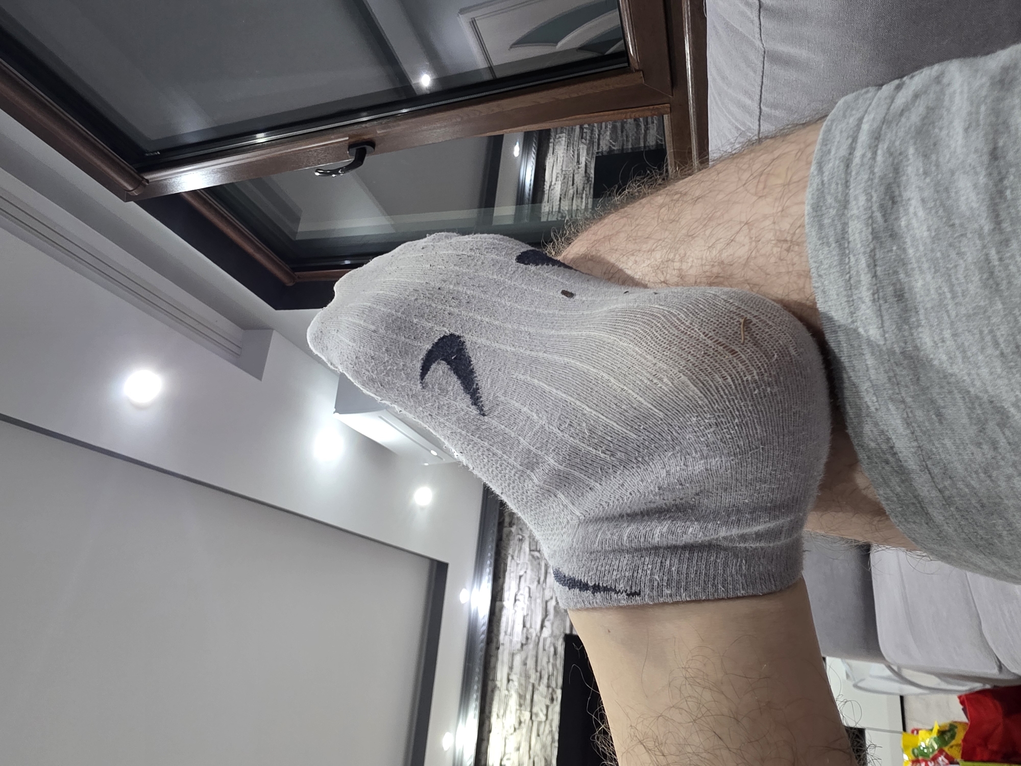The image depicts an indoor scene in Yagodovo, Bulgaria, viewed from a low angle, focusing on a person's elevated lower legs and the surrounding environment. The primary subject is a hairy leg, wearing a light grey, ribbed athletic sock with a dark grey or black swoosh-like logo visible on the top of the foot. The sock exhibits signs of wear and dirt, particularly around the heel. The foot is elevated, suggesting a resting posture, with the leg possibly resting on the person's other leg or a piece of furniture. The person is wearing light grey athletic-style shorts, visible on the lower right.
The background reveals a contemporary living space. To the right, a large, dark-framed window, possibly double-glazed, extends upwards, showing a handle and reflecting elements of the room's interior, including a white door with a distinct curvilinear panel design and a portion of the textured wall. Above the window, a white ceiling features a recessed section and multiple bright, circular LED spotlights. To the left and center-rear, a feature wall constructed from light-colored, stacked stone or brick veneer is visible, with a dark, rectangular recess below, possibly indicating a fireplace. Additional ceiling lights are scattered across the white ceiling. A grey upholstered sofa or couch is partially visible in the lower foreground and lower right, indicated by its material and stitching. In the extreme lower-left corner, a red bag and a colorful, crinkled package, possibly containing snacks, are partially visible on what appears to be the floor or a low surface. The overall lighting is artificial and bright.