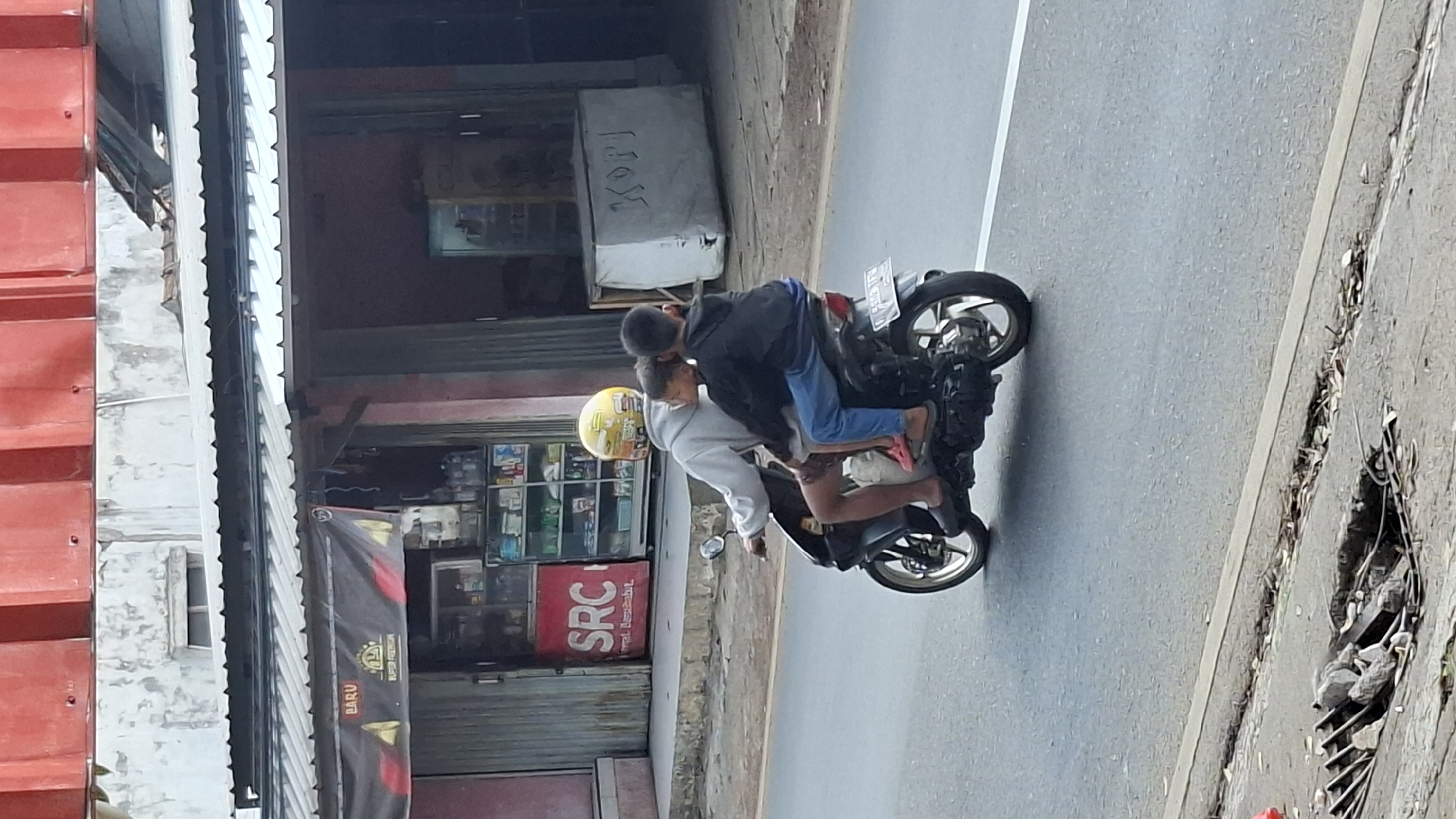In Cilaku, Indonesia, three individuals are riding on a black motorcycle traveling on an asphalt road. The person in the front is wearing a light gray hoodie and a yellow helmet with a sticker on it. They are driving the motorcycle, and their legs are visible, wearing dark shorts and sandals. Behind the driver, two children are seated. The child in the middle is wearing a dark jacket and blue pants, and their head is turned towards the child behind them. The child in the back is wearing a dark jacket and appears to be leaning on the child in the middle. The background shows a building with a red corrugated roof and a gray concrete wall with peeling paint. Below the roof, a shopfront with various shelves of goods is visible. A black banner with the word "BARU" and a logo is displayed above the shop entrance. To the right of the shop, a large, white, rectangular object with the word "KOPI" written in black letters is visible. The road in the foreground has a concrete curb and a partially visible drainage grate filled with debris.