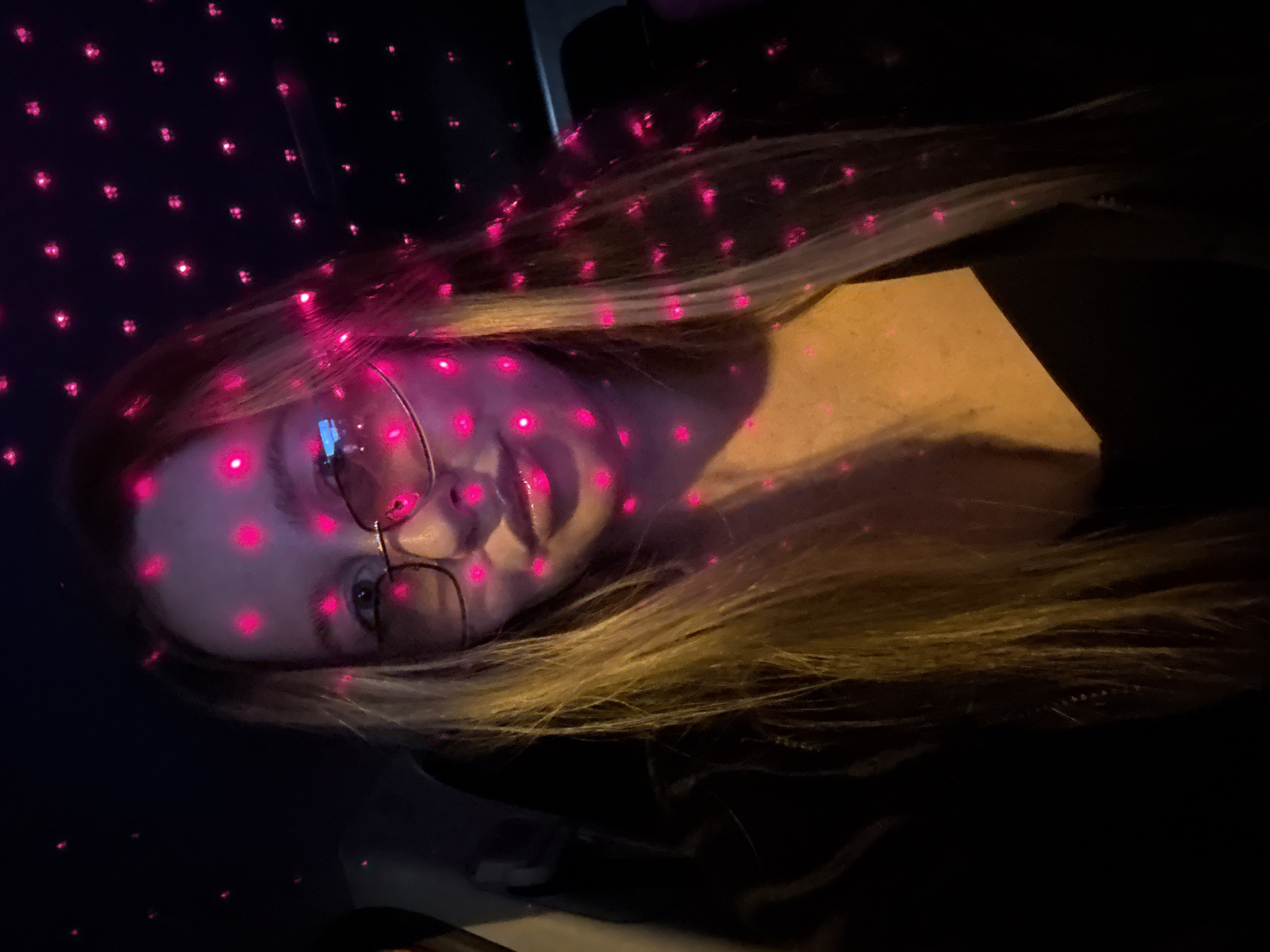 A young woman with long blonde hair and glasses is in the back seat of a car, illuminated by pink starry lights projected onto the interior. The lights create a bokeh effect across her face and hair. She is looking directly at the camera with a neutral expression. The background is dark, suggesting it is nighttime or the interior of the car is dimly lit, with the exception of the pink light projections. The scene appears to be taking place in the city of Chaikovsky, Russia, although there are no specific visual cues in the image to confirm this location.