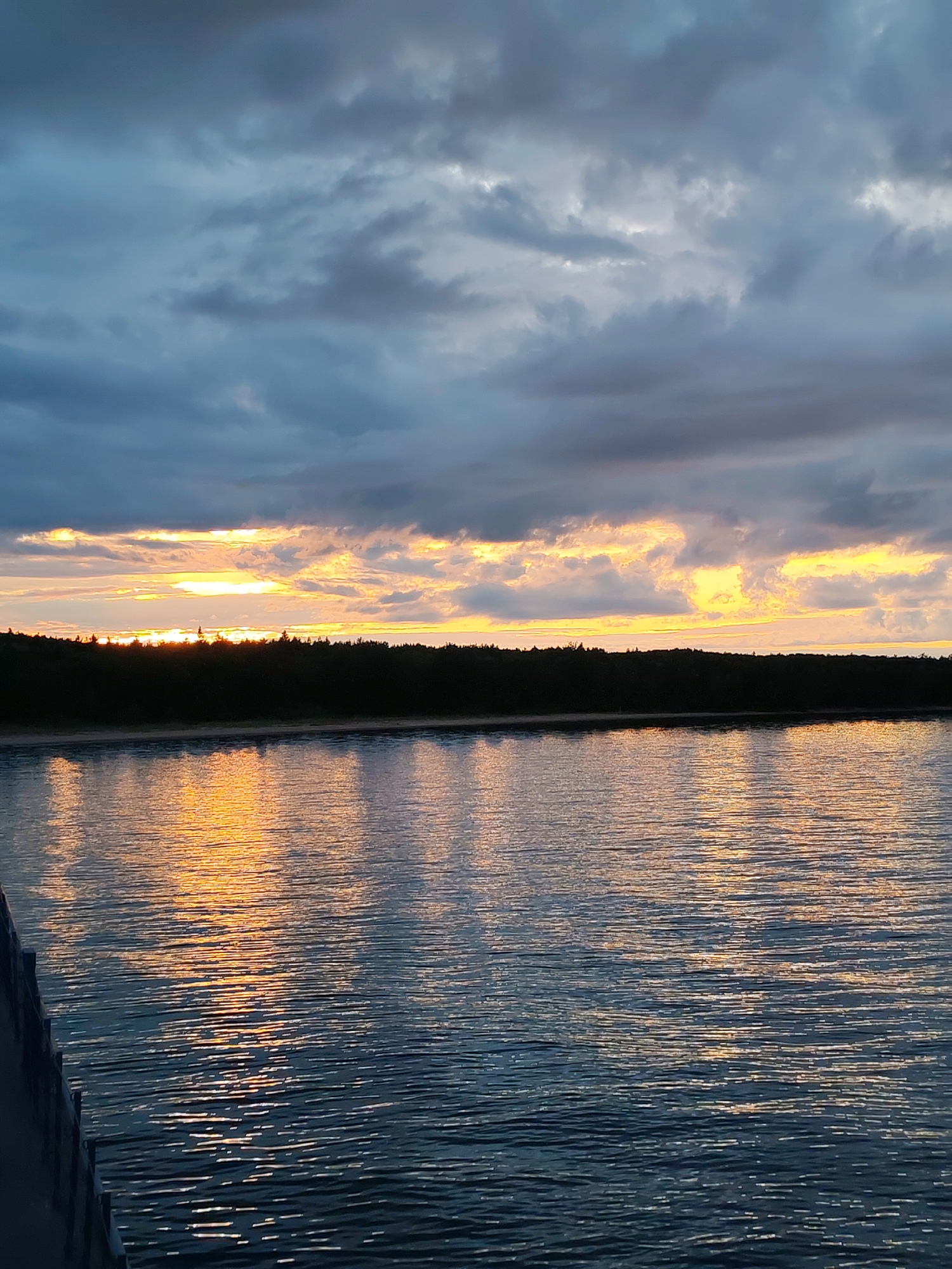 The image captures a sunset over a body of water, possibly a lake or a wide river, in the village of Shcheleiki, Russia. A dark, treed shoreline separates the water from the sky. The sky is filled with dramatic, heavy clouds, tinged with orange and yellow where the sun is setting below the horizon. The setting sun casts a bright, golden reflection on the rippling surface of the water. In the foreground, the edge of a dark structure, likely a pier or boardwalk, is visible on the left side of the frame. There are no people or animals visible in the scene. The overall atmosphere is serene and perhaps a little moody due to the dark clouds.