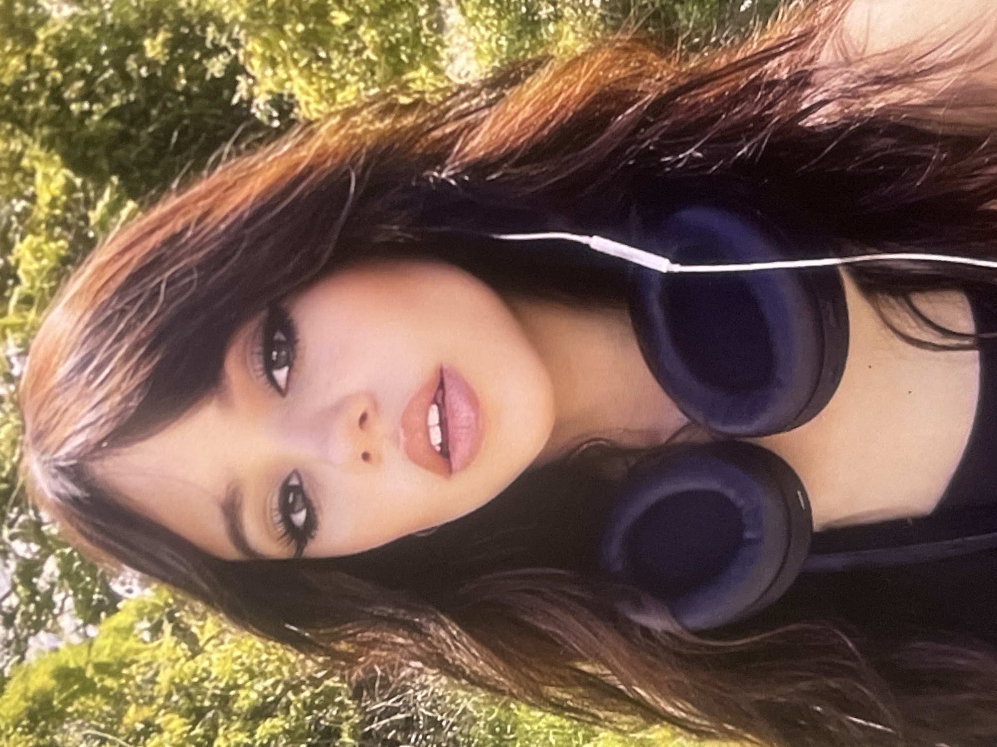 A close-up, informal portrait of a young woman, likely taken outdoors in Poltava, Ukraine, during the daytime. The woman has long, dark brown hair with noticeable auburn highlights, framing her face and flowing over her shoulders. She wears prominent eye makeup, featuring extended eyeliner and dark, full lashes that accentuate her dark eyes. Her eyebrows are well-defined. Her lips are slightly parted, revealing her upper teeth, and are colored with a natural or light pink/peach hue. A small dark mole is visible on her right cheek, near the corner of her mouth. Around her neck rest a pair of dark blue, over-ear headphones, connected by a white audio cable that runs across her chest. A small white "L" marking is visible on the left ear cup. The background is a soft blur of vibrant green foliage, suggesting a sunny, natural outdoor setting such as a park or garden. The woman maintains a calm and composed expression, looking directly at the camera.