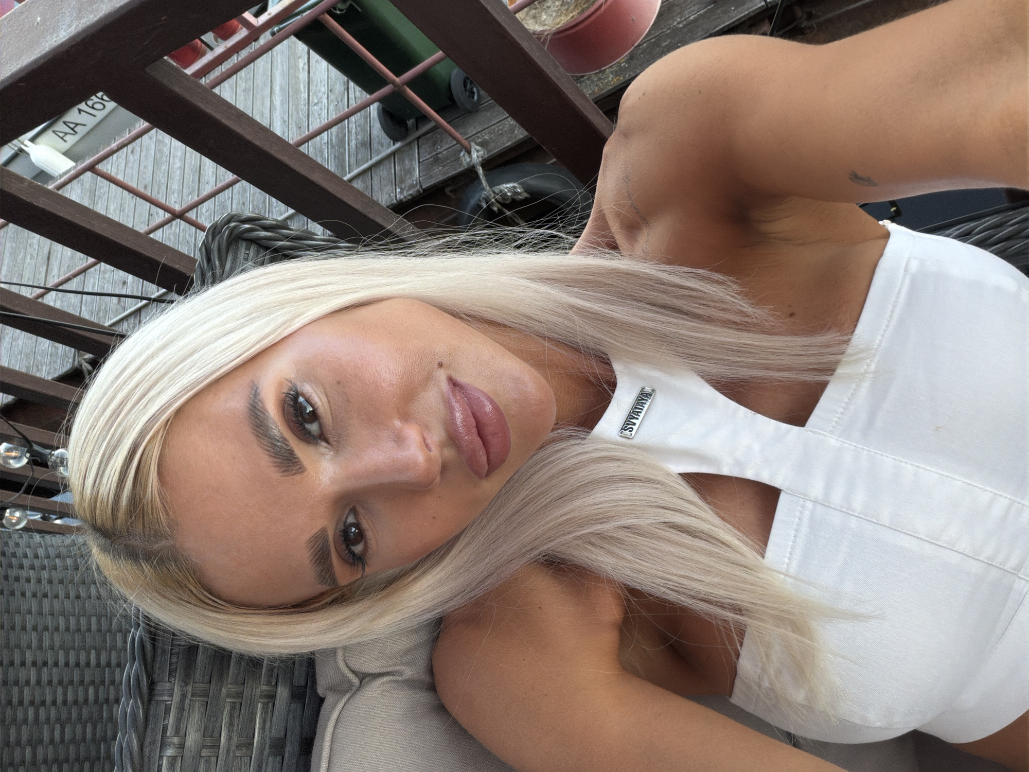 A young Caucasian woman with blonde hair is taking a selfie outdoors. She is wearing a white top. The background includes a railing and wooden decking, suggestive of a balcony or pier. A sign with "AA 166" is visible in the background. The lighting indicates it is daytime. The scene is not identifiable as being in Kazan, Russia.