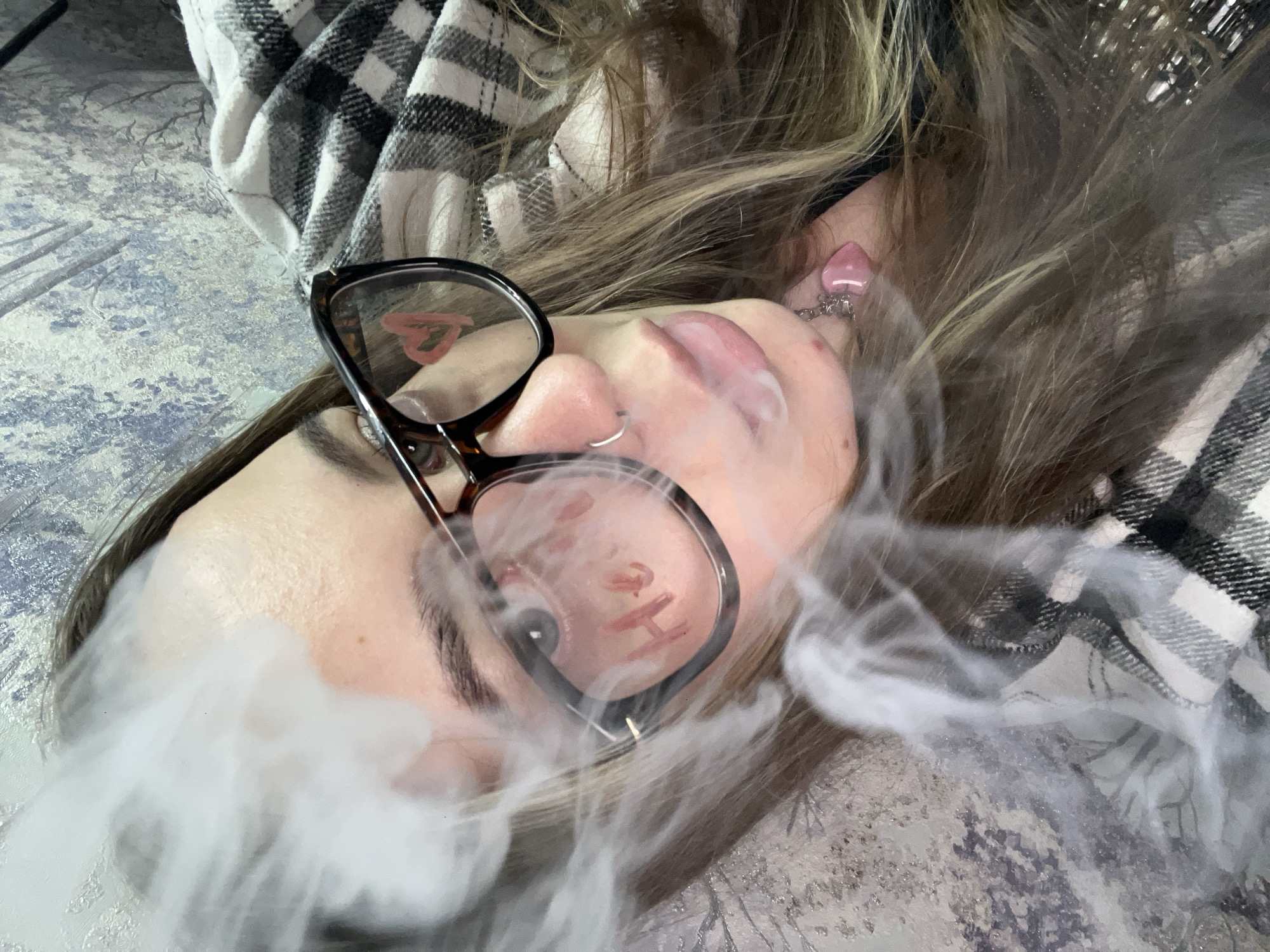 The image shows a person lying down, exhaling a cloud of vapor. The person is wearing glasses, which reflect an image, and has blonde hair and piercings in their nose and ear. They are lying on a surface with a patterned fabric. The location, Novopokrovskaya, Russia, is not visually discernible from the image.