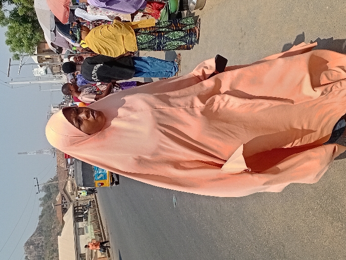 A woman in an orange hijab stands on a roadside, facing the camera. Behind her, people and stalls suggest a market scene. The background shows a landscape with buildings and a hill. The woman's expression is neutral, and the hijab covers her head and body, with a section on the ground. The setting appears to be a bustling outdoor environment with varied activities.