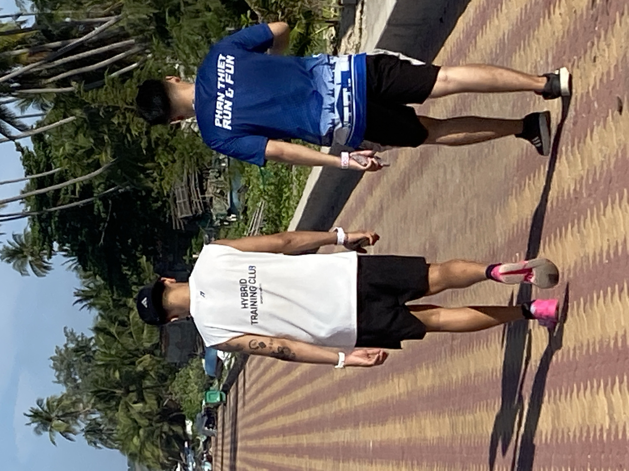 Two individuals, viewed from behind, are walking on a paved path with a patterned surface of yellow and red stripes. The person on the left wears a white sleeveless shirt with blue text that reads "HYBRID TRAINING CLUB" and black shorts. They have tattoos on their left arm and wear a black baseball cap, a white wristband on their left wrist, and pink and grey athletic shoes. The person on the right wears a blue t-shirt with white text that reads "PHAN THIET RUN & FUN" and a white and blue graphic design along the bottom. They also wear black shorts and black athletic shoes. Both individuals have white wristbands on their right wrists.
The background features a vibrant tropical environment. Lush green palm trees and other foliage line the right side of the path, with tall palm trees in the distance. Behind the foliage, parts of buildings and structures, some appearing weathered or under construction, are visible. Boats are also present near the shoreline, suggesting a coastal or near-water location. The path appears to be an elevated walkway or promenade, with a low concrete barrier on the right side separating it from the background. The lighting suggests it is daytime, with clear skies and shadows cast on the path. The location is Mũi Né, Vietnam, a coastal town known for its beaches and watersports.