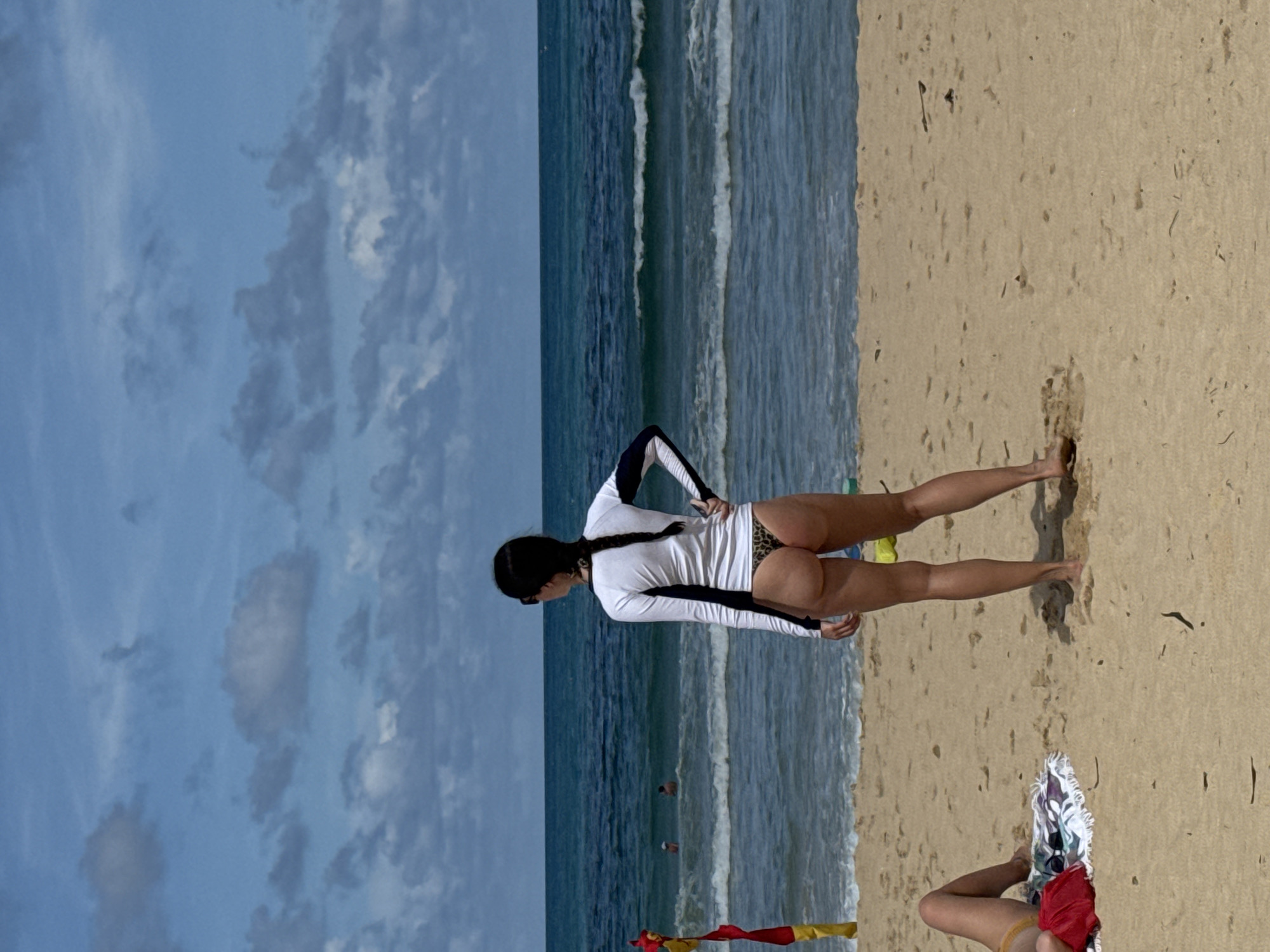 In Rawai, Thailand, a woman stands on a sandy beach, facing the ocean. She is wearing a white long-sleeved rash guard with dark blue trim and a leopard-print thong. Her hair is braided and falls down her back. She is wearing sunglasses and has her hand on her hip. In the background, the ocean has waves, and a few people are in the water. To the left of the frame, a red and yellow flag is planted in the sand. In the foreground on the left, another person's legs are visible, lying on a towel. The sky is blue with scattered clouds.