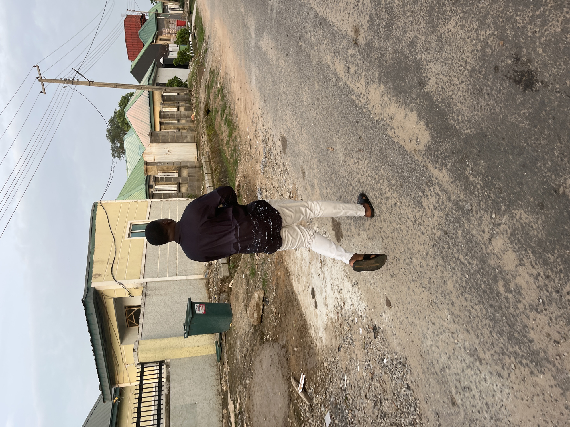 In Abuja, Nigeria, a man walks away from the camera down a dirt and asphalt road. He is wearing a dark, long-sleeved shirt with a speckled pattern on the bottom half and white pants. He has on dark sandals. To his left, there is a green trash bin next to a yellow and gray building with a black metal fence. In the background, there are several small, single-story houses with green roofs. A wooden utility pole with multiple wires runs along the right side of the road. The sky is overcast.