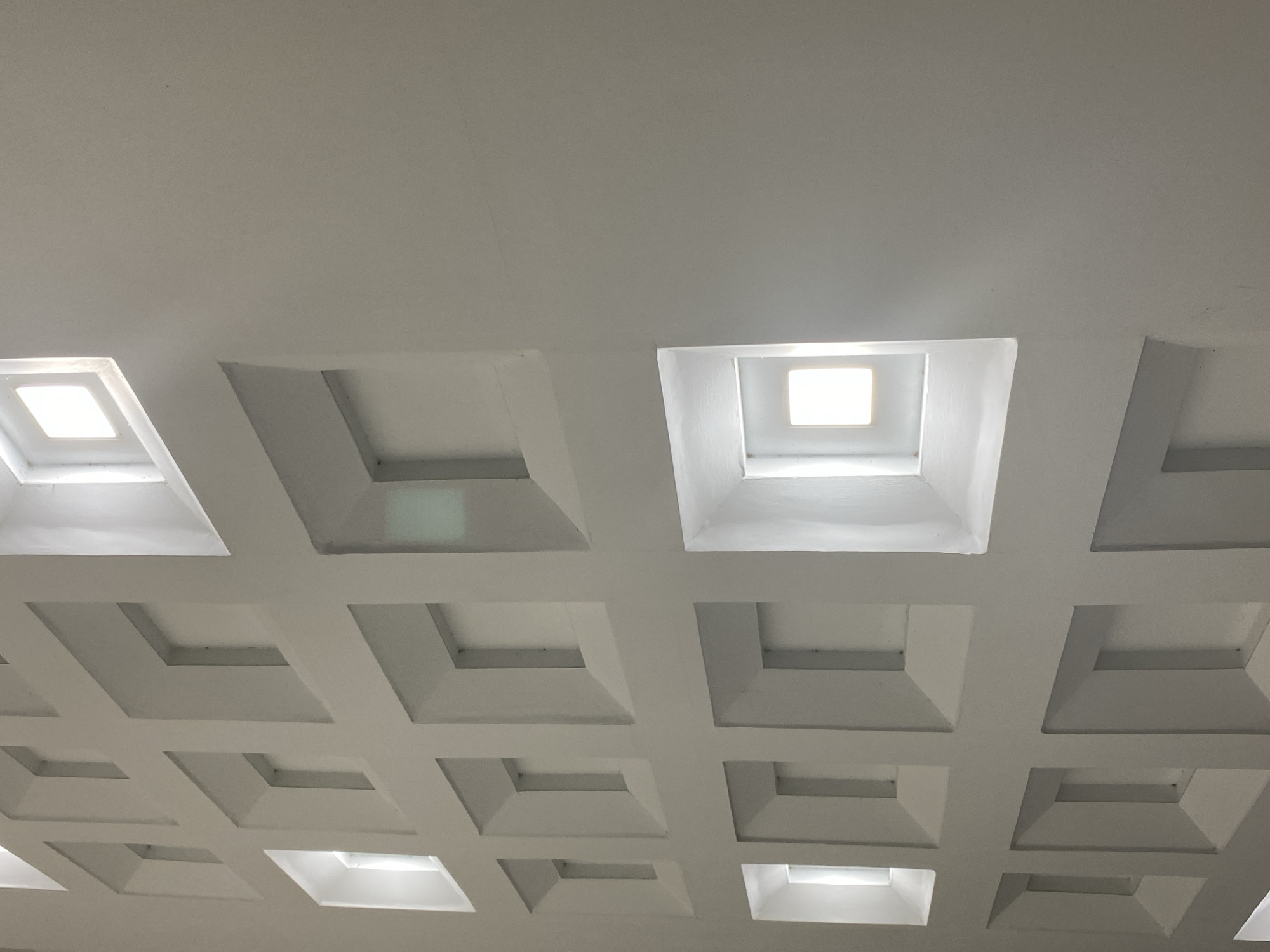 The image shows a coffered ceiling with recessed square light fixtures. The ceiling is white and appears to be made of plaster or a similar material. The lighting fixtures are arranged in a grid pattern. Some of the fixtures are illuminated, while others are not. The perspective is looking up at the ceiling from below. The specific location in Banda Aceh, Indonesia, is not discernible from the image.