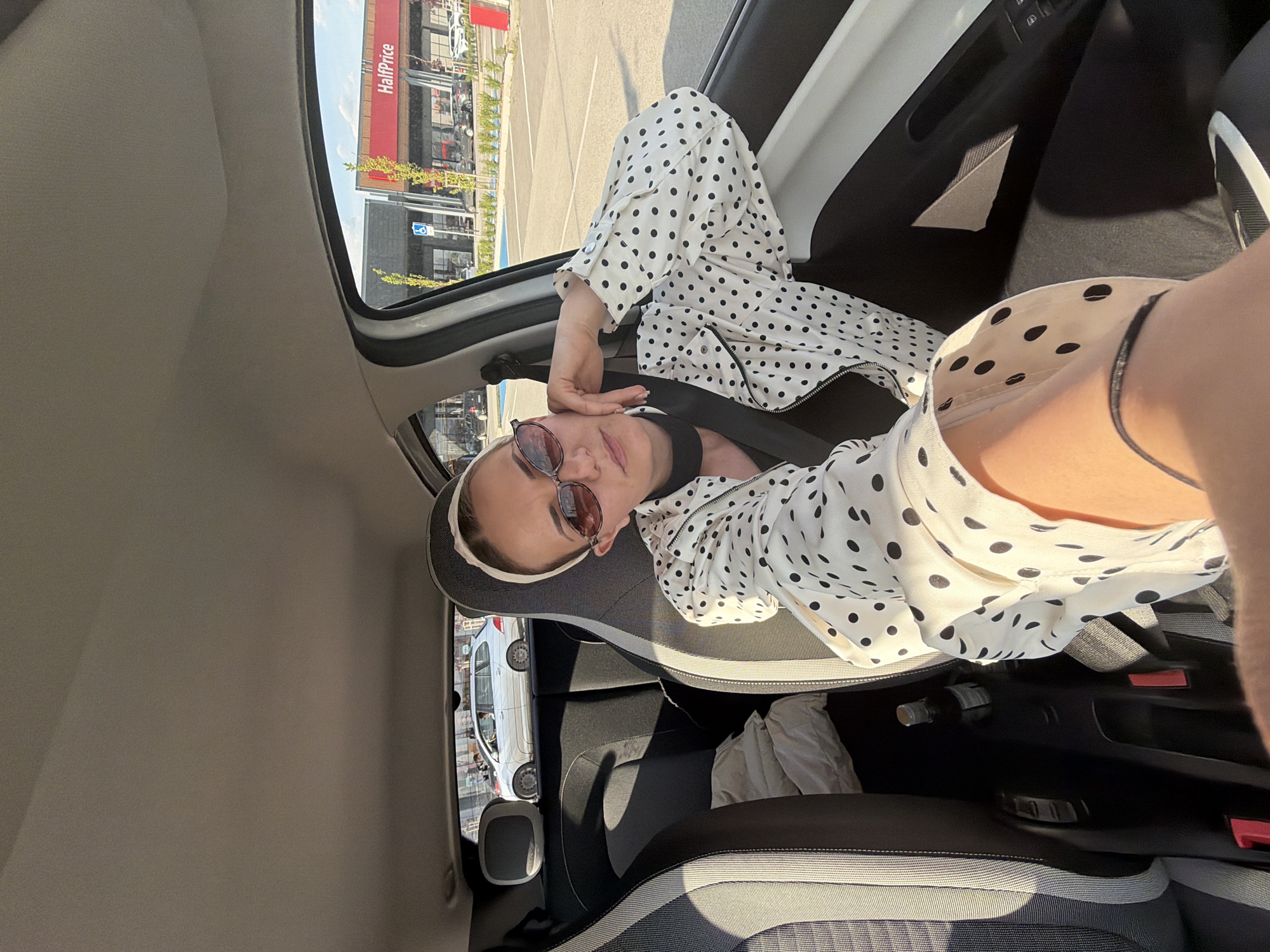 The image shows a woman sitting in the passenger seat of a car. She is wearing a white jacket with black polka dots, sunglasses, and a white headband. She is wearing a seatbelt. Through the open car door, a building with a red awning bearing the name "HalfPrice" is visible, suggesting the location is likely near a shopping area. The image was taken during daylight, as indicated by the bright sunlight and clear sky visible through the car window. Based on the architectural style and the "HalfPrice" store, the location is identified to be near Sofia, Bulgaria. There is an open bottle or container in the center console. There's a crumpled, possibly white, item on the backseat behind the passenger seat.