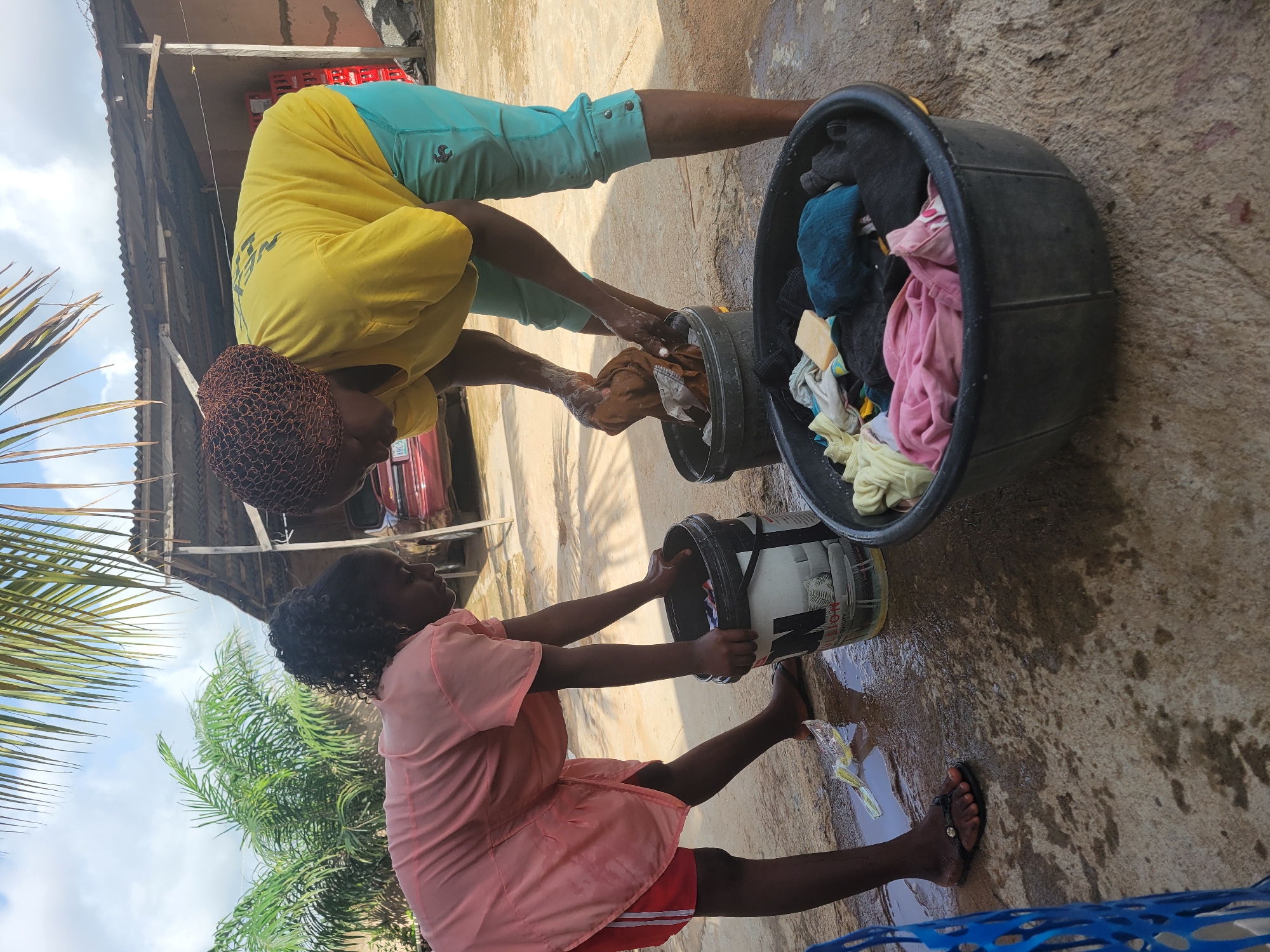 Two women are engaged in washing clothes outdoors in what appears to be Madalla, Nigeria. One woman, wearing a yellow shirt and teal capri pants with a net cap on her head, is bent over a gray bucket, washing clothes. The other woman, in a pink top and red shorts, is similarly positioned over a white bucket with black lettering. A large, dark gray basin filled with clothes sits nearby. The ground is wet, and the surrounding environment includes a corrugated roof, palm trees, and a glimpse of a red vehicle.