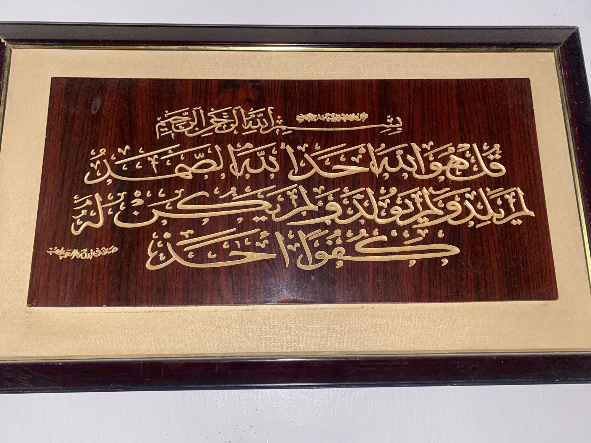 This media file is a close-up, eye-level shot of a framed piece of Islamic calligraphy. The artwork is mounted on a dark, polished wood plaque and framed with a dark brown border. The calligraphy itself is in a golden, raised font, set against the rich, dark wood. The inscription appears to be from the Quran, with the top line including "بِسْمِ ٱللَّٰهِ ٱلرَّحْمَٰنِ ٱلرَّحِيمِ" (In the name of Allah, the Most Gracious, the Most Merciful). Below this, larger Arabic script is visible, followed by a smaller inscription in the bottom left corner which reads "صدق الله العظيم" (Allah the Almighty has spoken the truth). The overall impression is one of religious devotion and artistic craftsmanship. The lighting is even, suggesting indoor display. No specific time of day, weather, or location cues are visible beyond the context of Fagge A, Nigeria.
