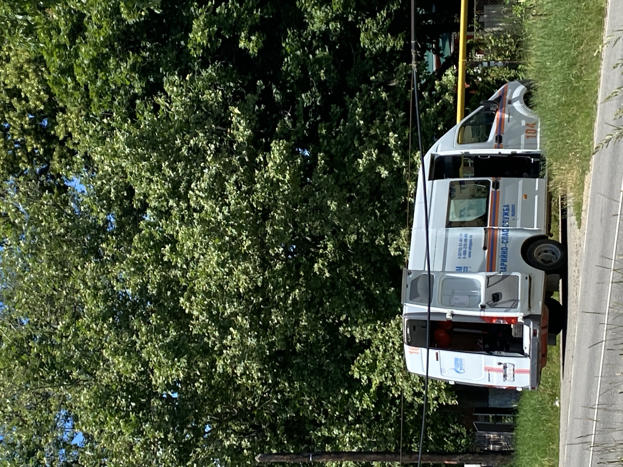 A white van with emergency markings is parked on the side of a road in what appears to be a suburban or rural area of Zapadny, Russia. The van's rear doors and the side passenger door are open, revealing equipment and personnel inside. The van has the logo of "GAZPROM" and what appears to be emergency service markings in Russian. A large, leafy tree dominates the background, with a wooden utility pole and wires in the foreground. The weather is sunny, and the vegetation is green and lush, suggesting a warm season.