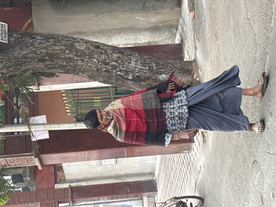 An elderly man walks barefoot on a paved path. He wears a plaid shawl draped over a checkered shirt and blue, draped trousers. A large tree trunk is visible in the background along with a building and its fence. A bicycle lies on the path near the man. A small sign is affixed to the tree.