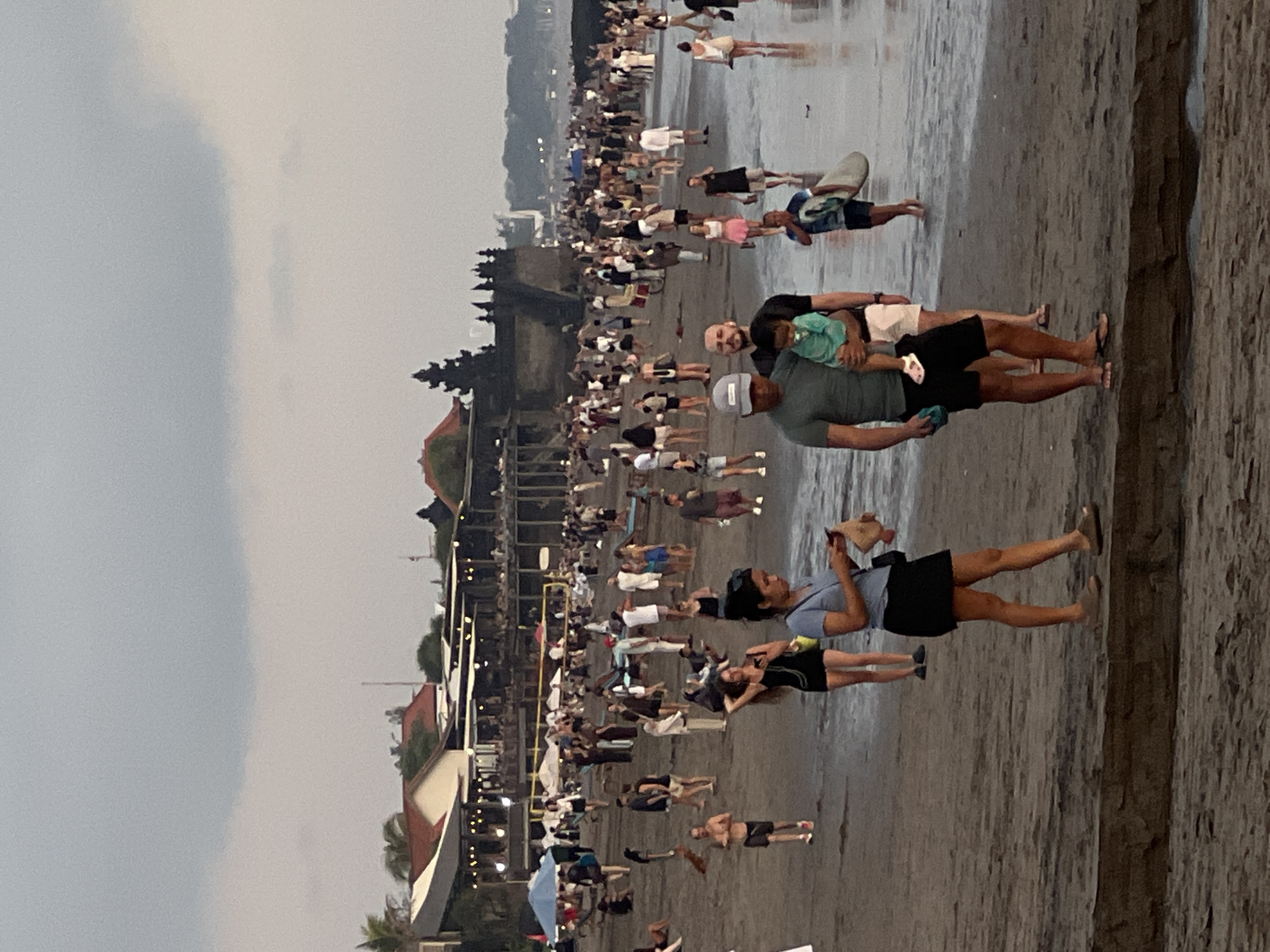 The image captures a crowded beach scene in Canggu, Indonesia, with numerous people milling along the shore. The sky is overcast, contributing to a muted color palette. The beach is wet, reflecting the figures and structures. A temple complex with multi-tiered structures is visible in the background. Close to the viewer, people are standing, walking, and looking towards the ocean. To the far right, there is what appears to be a retaining wall.