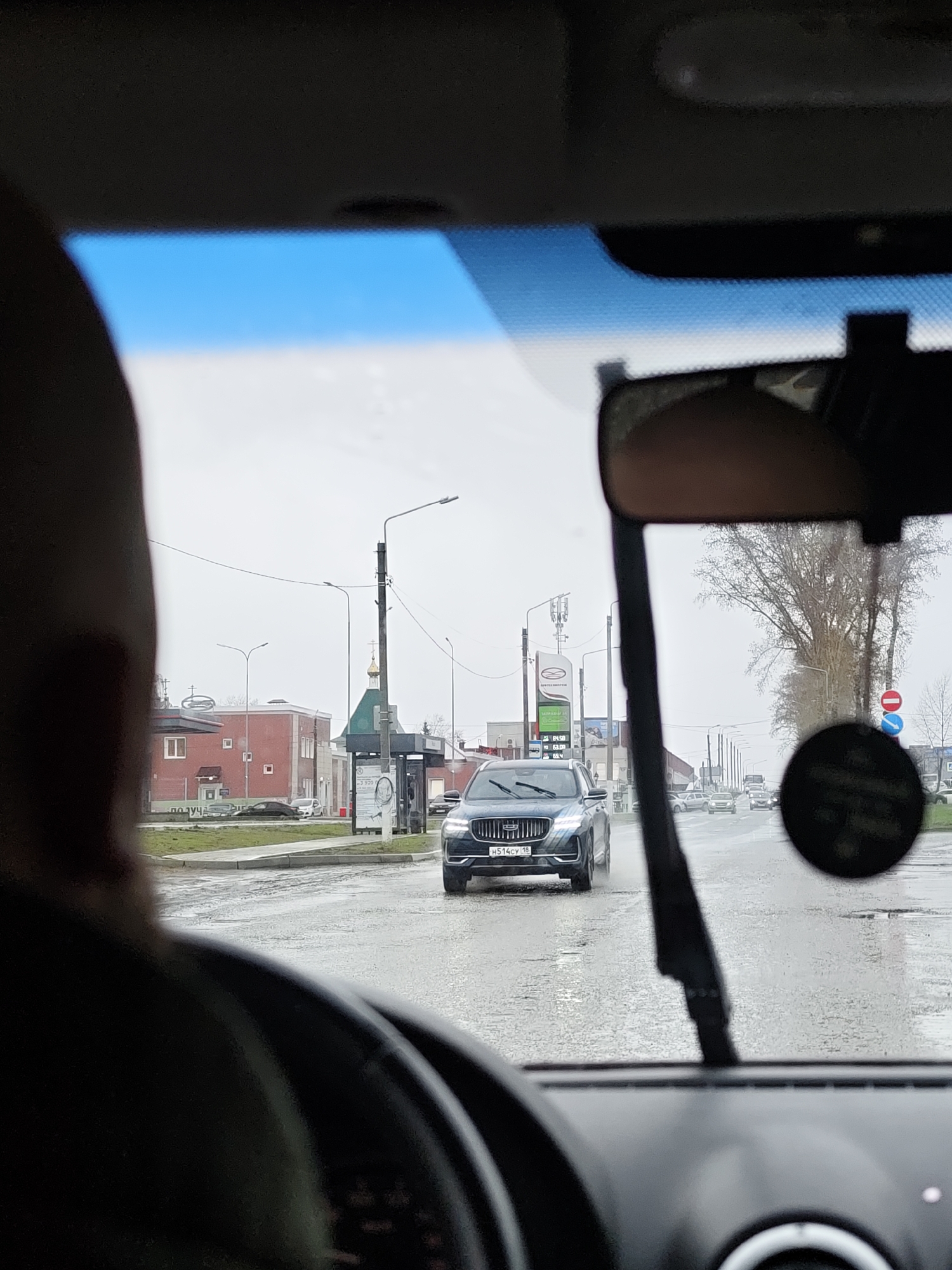 The image captures a first-person perspective from inside a car, looking out through the windshield at a street scene in Chaikovsky, Russia. The foreground shows the driver's left shoulder and head, partially obscured, along with the steering wheel and dashboard. A sun visor is visible at the top, and the rearview mirror on the right frames part of the scene.
The weather appears overcast, and the road is wet, suggesting recent rain or drizzle. Approaching in the center of the frame is a dark grey SUV, possibly a Geely, displaying a Russian license plate "H514CV 18". The "18" indicates the Udmurt Republic region.
To the left of the road, various buildings line the street. A red-brick building has small square windows. Further back, a gas station sign with Cyrillic text "ОПТАН" (Optan) and fuel types "Дизель", "АИ-92", "АИ-95" with prices is visible. Another sign behind it reads "СТОМАТОЛОГИЯ" (Dentistry). A bus stop with an advertisement, partially showing "ПОЛУЧИ" (receive/get), is also present. A distinctive golden onion dome of an Orthodox church can be seen peaking above some structures. Streetlights and power lines crisscross the sky. To the right, trees with bare branches line the road, and a red circular "no entry" sign is visible. Other vehicles are present further down the road.