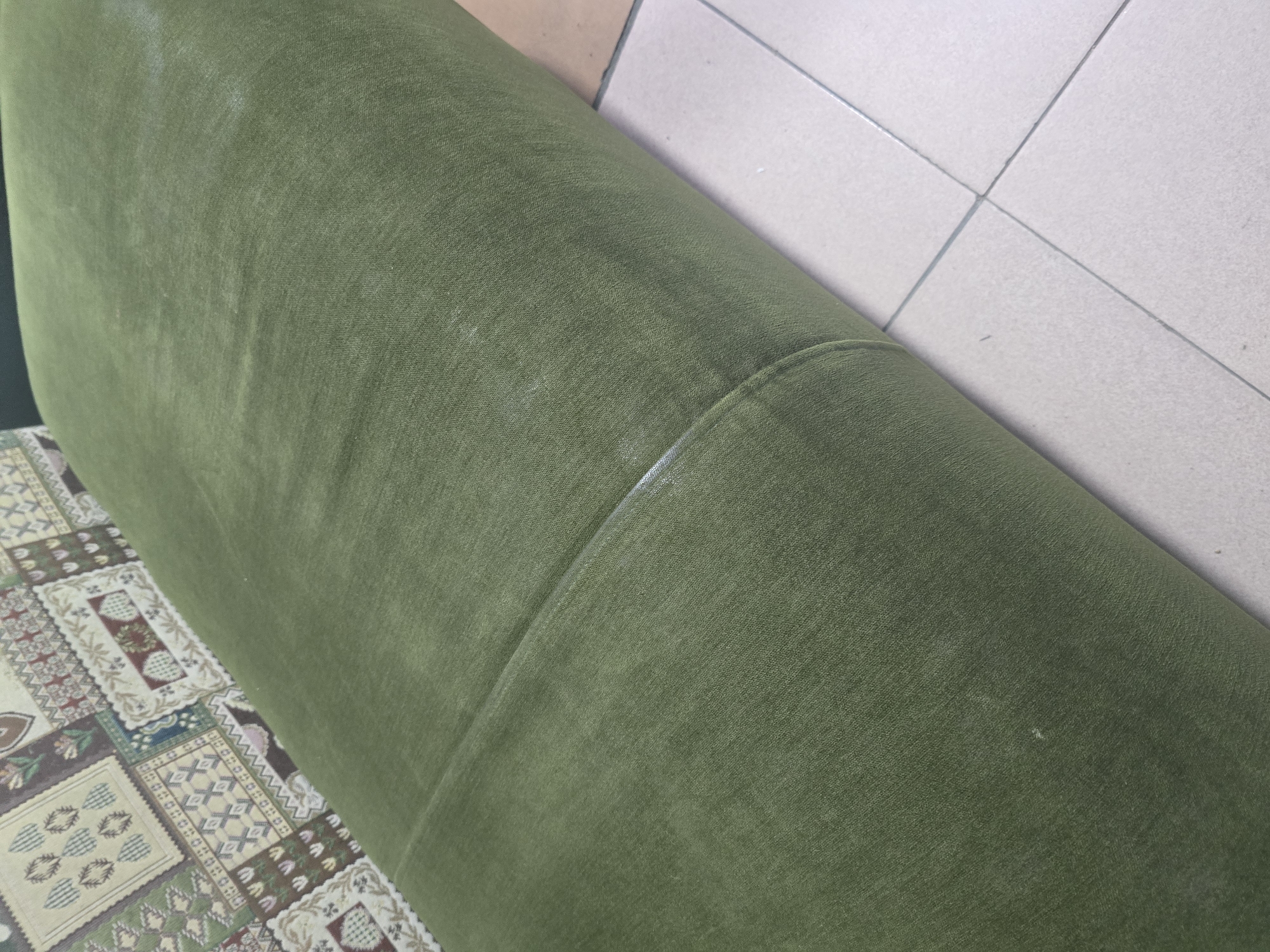 A close-up shot shows a rolled-up green carpet with a patterned fabric laid underneath. The patterned fabric is visible on the left side of the image, featuring a quilt-like design with floral and geometric motifs in various colors. The carpet is a solid dark green color and appears to be made of a soft material. It is rolled from right to left, with the edge of the carpet visible. The floor is tiled with light-colored squares.