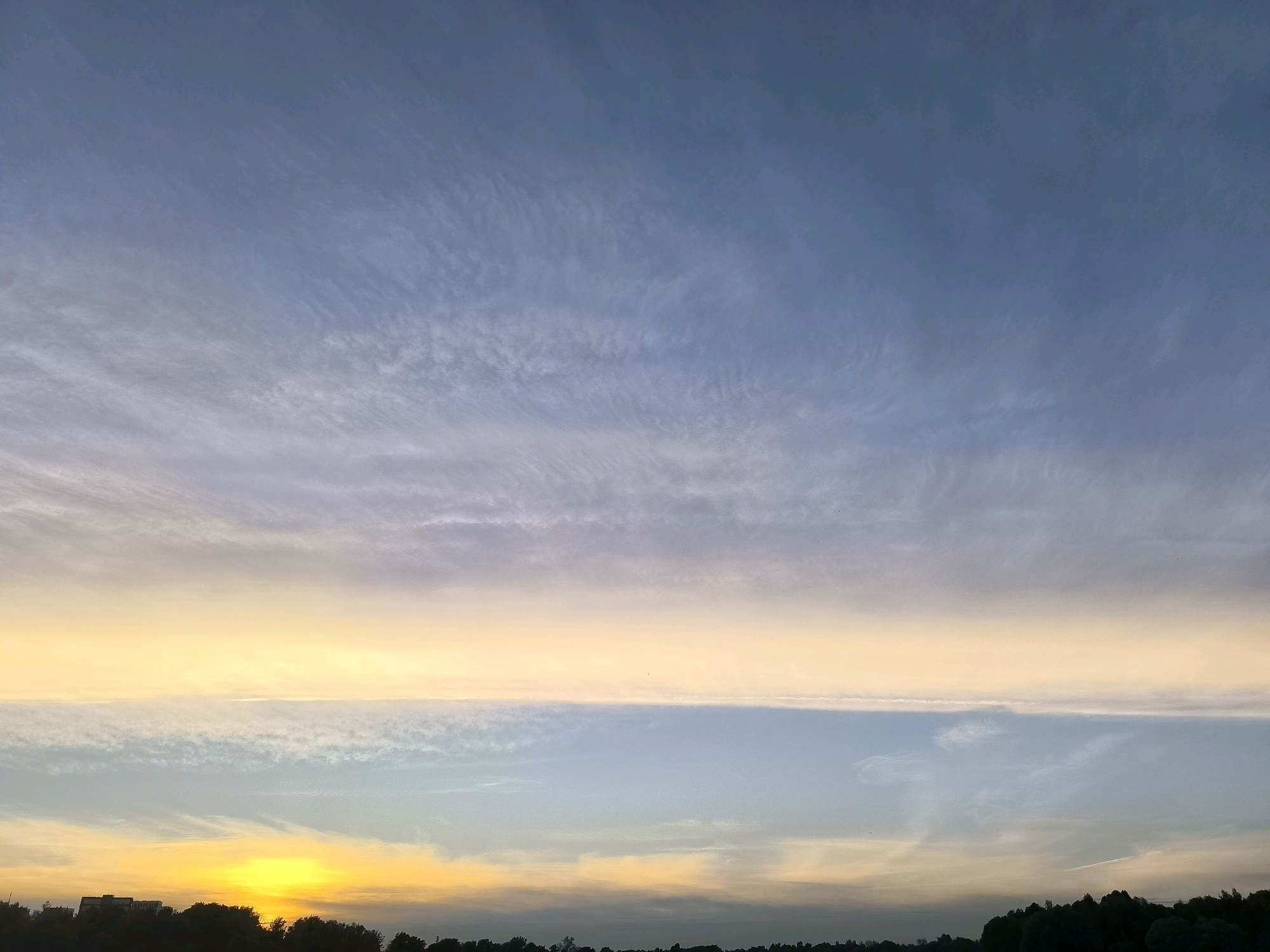 The image depicts a sunset over the city of Dolgoprudny, Russia. The sky is filled with streaks of clouds, colored in hues of blue, yellow, and orange. The sun is setting on the left side of the image, casting a bright yellow glow. The lower part of the image shows a dark silhouette of trees and buildings against the colorful sky. There are no people or animals visible in the image. The scene is a natural landscape illuminated by the setting sun.