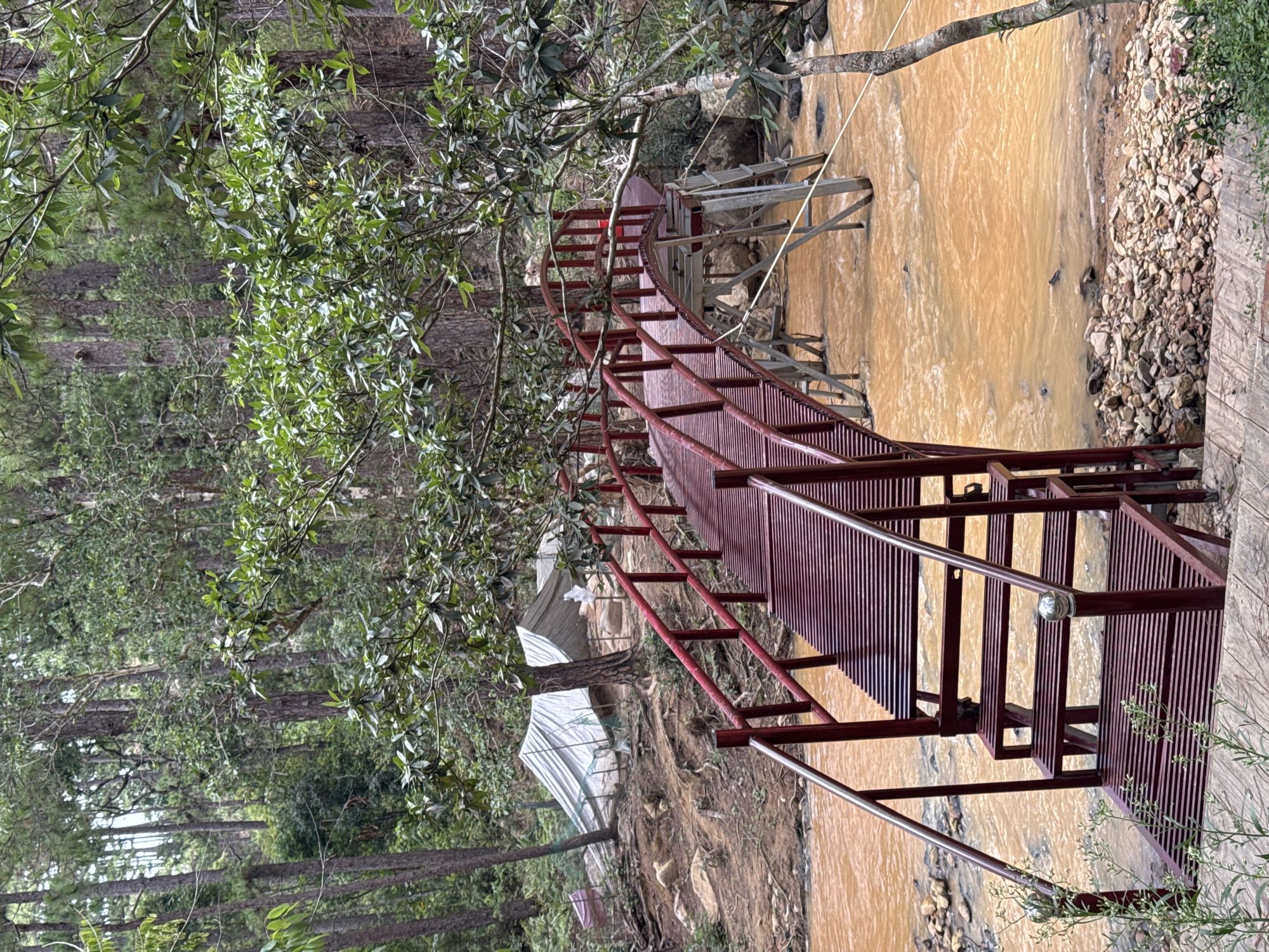 The image shows a metal pedestrian bridge spanning a murky, brown river or stream. The bridge is painted red and has a curved design with steps leading up to it from one side. The surrounding environment is lush with greenery, including trees with dense foliage. In the background, there are structures that appear to be greenhouses or covered agricultural areas. The setting is likely a rural or semi-rural area, possibly near the city of Lang Biang - Đà Lạt, Vietnam, known for its agriculture and natural landscapes. The absence of people suggests a tranquil and possibly remote location.