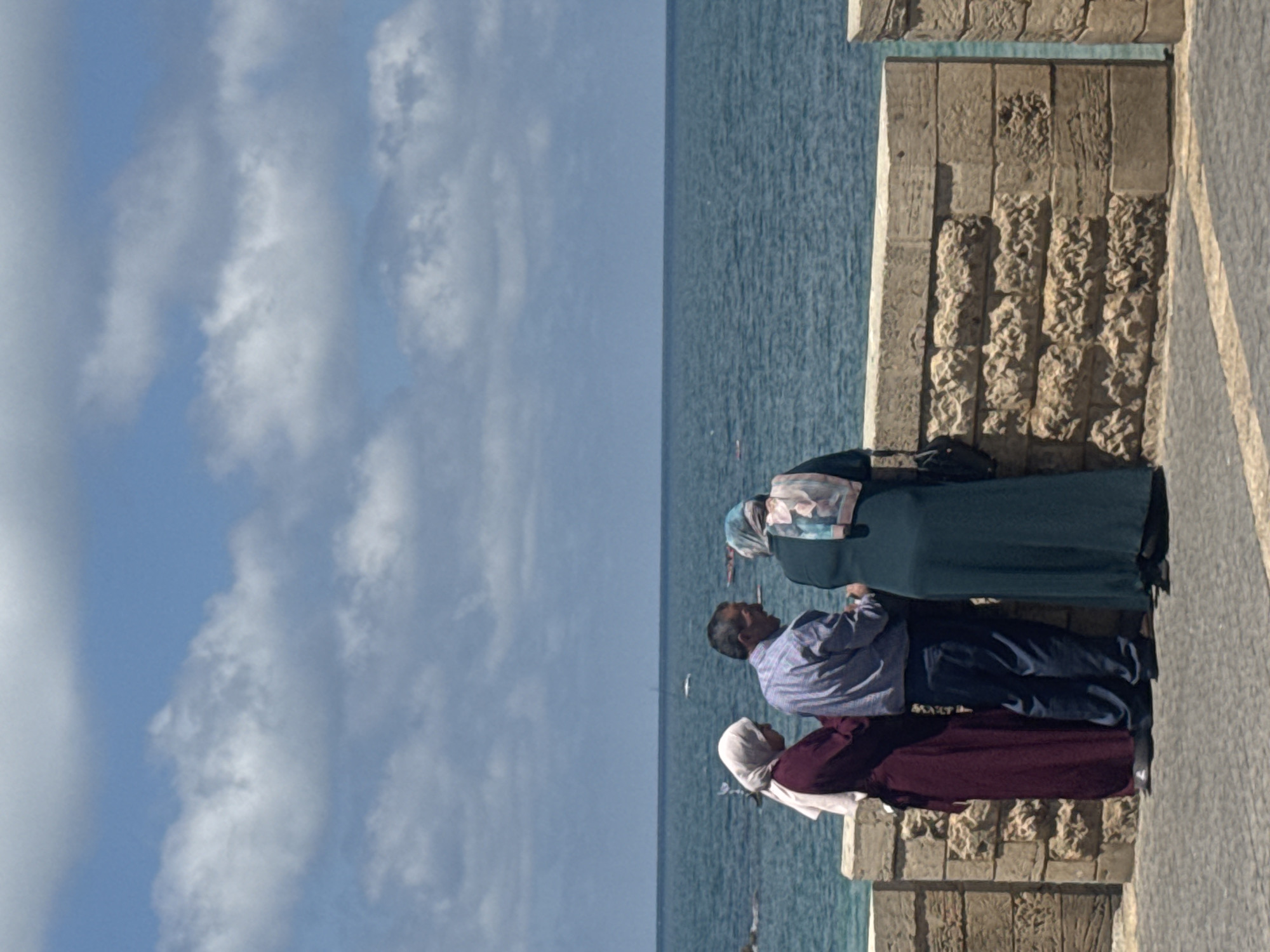 Here is a description of the image:
Three individuals are standing near a stone wall that appears to be overlooking the sea. The sky is visible in the background and features scattered clouds. The person closest to the camera is wearing a white head covering and a maroon-colored outfit. Next to this individual is a person wearing a blue and white checkered shirt with dark pants. The third person is wearing a light-colored head covering with a long, dark garment.
Based on the environment and architecture, it's plausible that the location is Tel Aviv, Israel, as the coastal city features a blend of modern and historical stone structures along its Mediterranean coastline. The people are likely tourists.