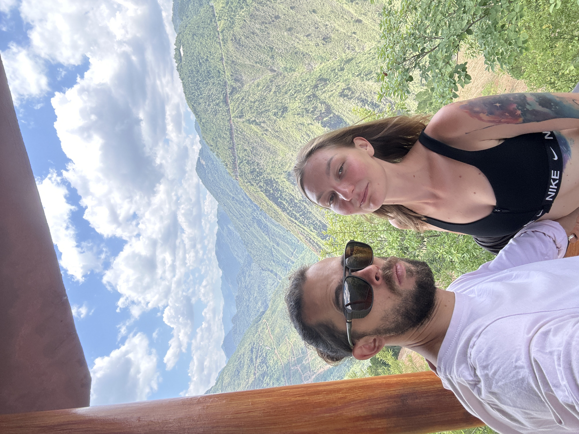 Two people are in a selfie photograph with a scenic mountain background. The woman is fair-skinned, wearing a black sports bra with "NIKE" visible, and has a tattoo on her upper arm. The man has a beard, sunglasses, and is wearing a white t-shirt. They appear to be on a balcony or platform with a wooden railing. The background features a steep, verdant mountain landscape under a partly cloudy sky. The location is Lũng Cú Commune, Vietnam, indicated by the mountainous landscape typical of the region.