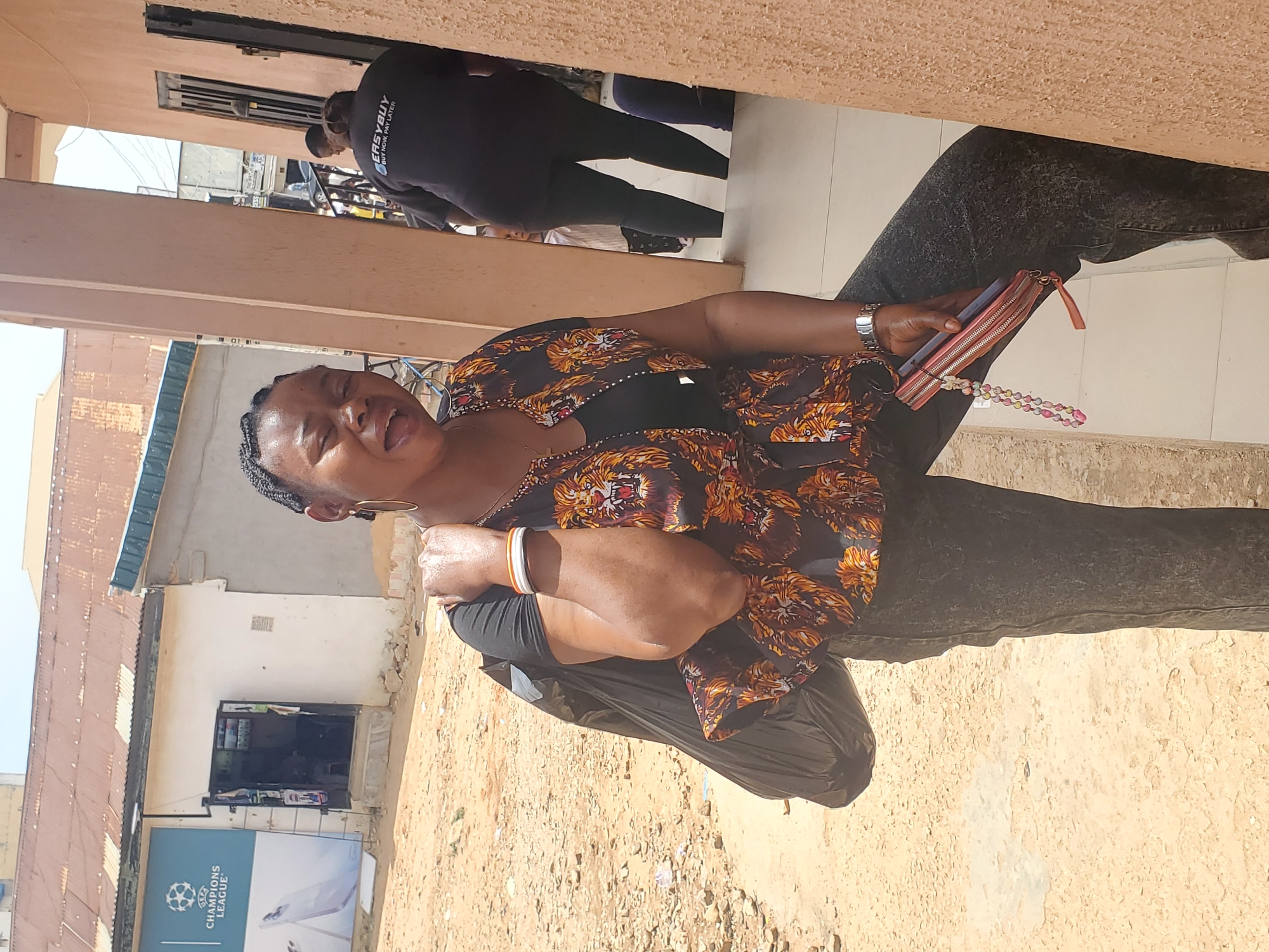 This image, set in Jos, Nigeria, captures an outdoor scene during the bright daytime. The primary subject is a woman with dark skin and braided hair, positioned in the foreground. She is dressed in a black and orange/gold patterned top over a black undershirt, dark-colored jeans, a wristwatch on her left wrist, and white and orange bangles on her left forearm. She holds a reddish-pink zipped wallet with a colorful beaded strap in her right hand. Her facial expression, with an open mouth and squinted eyes, suggests she is experiencing a strong emotion, possibly laughter, shouting, or reacting to the bright sunlight.
The ground around her is composed of sandy dirt and some lighter-colored paved sections. In the background, other individuals are visible; one person, seen from behind, wears a black shirt with the text "EASYBUY BUY NOW. PAY LATER" clearly printed. Further back, buildings with simple architecture, including one with a corrugated metal roof, line what appears to be a street or commercial area. An open doorway on one building suggests a shop. A light blue banner is visible on a building wall, featuring the UEFA "CHAMPIONS LEAGUE" logo and text, indicating a local connection to football culture. The scene conveys a lively, everyday atmosphere in an urban or semi-urban Nigerian setting.