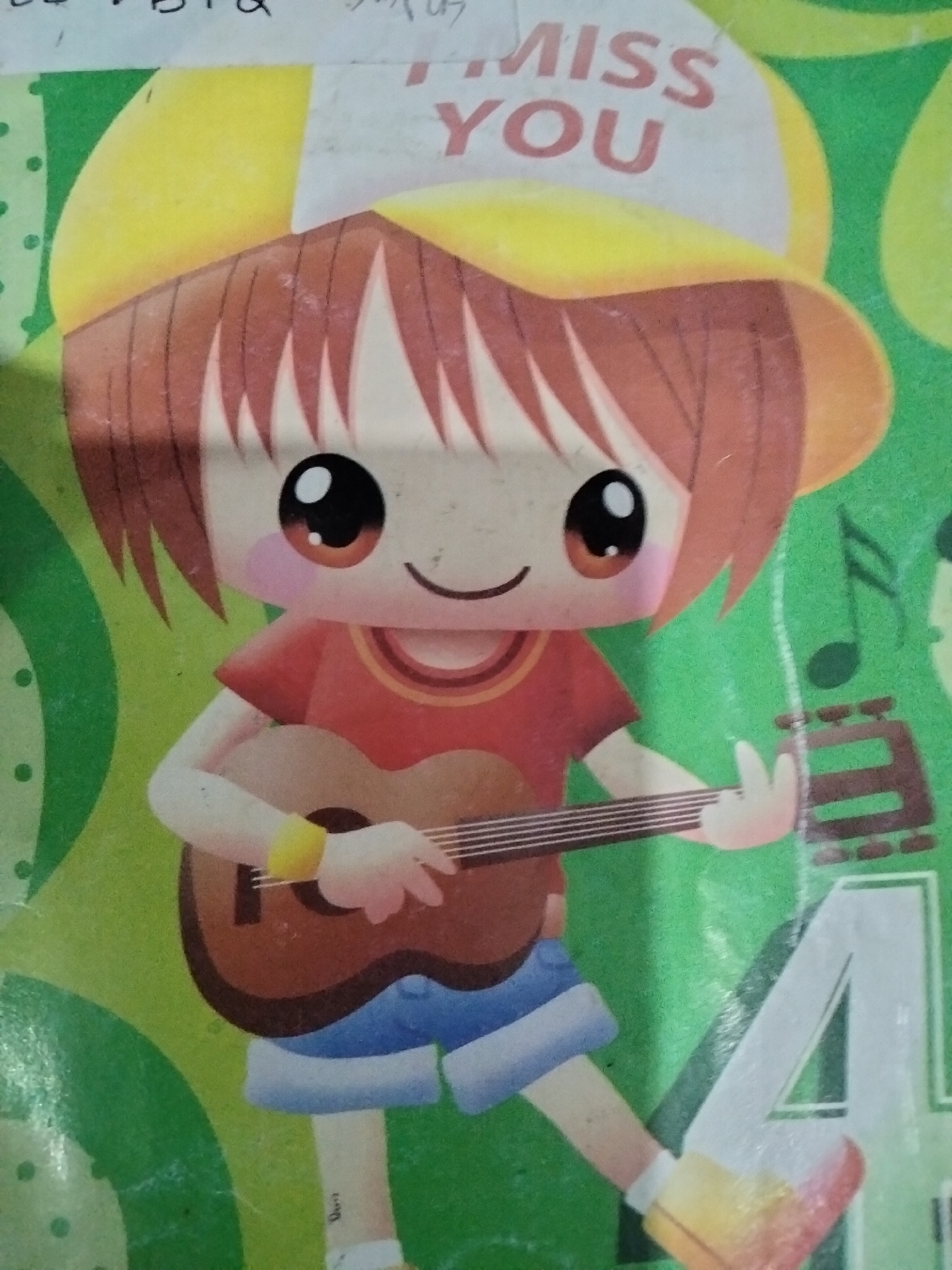 The image shows a cartoon illustration of a young person wearing a yellow cap and playing a guitar. The character has large, expressive eyes, red hair, and is dressed in a red shirt and blue shorts. The background is green with various patterns, including musical notes and a large white letter "A". There is text on the cap that reads "MISS YOU". The image appears to be a drawing or sticker. The location in the city of Payagaboh, Indonesia is not discernible from the image itself.