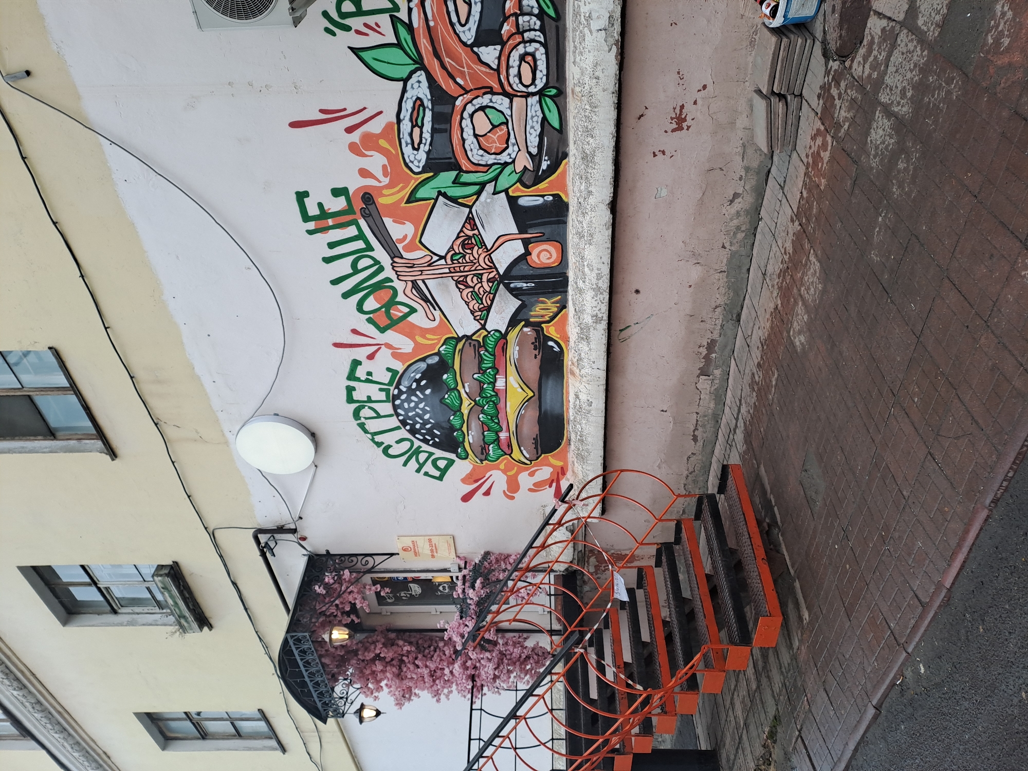 A daytime outdoor shot in Мининская Поляна Кп, Russia, features the side of a light yellow building with rectangular windows, adjacent to a wall adorned with a colorful mural. The mural includes sushi, a bowl of noodles with chopsticks, and a burger. Text is integrated into the design. A white, circular light fixture is visible near the mural. The wall is next to a sidewalk paved with reddish-brown bricks. A set of stairs with orange railings leads up to the building. Pink flowering branches hang near the entrance.