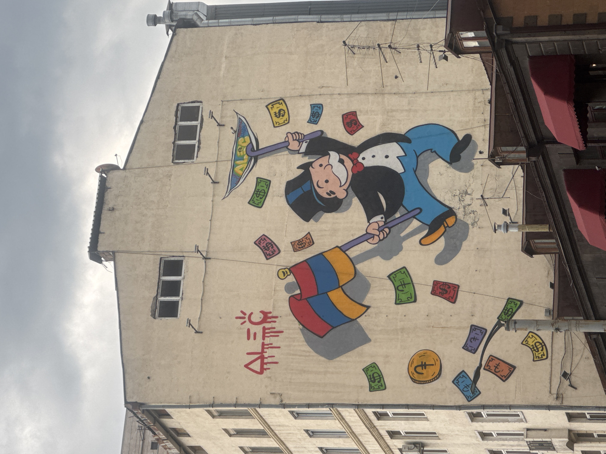 A large mural featuring the Monopoly man (Rich Uncle Pennybags) is painted on the cream-colored side of a tall building, viewed from a slightly upward angle under a cloudy sky. The cartoon figure, wearing a top hat, tuxedo, and red bow tie, is depicted in an active pose, holding a pickaxe marked with "¥€$" on its blade and a flag with horizontal red, blue, and yellow stripes (from bottom to top).
Numerous colorful cartoon banknotes, featuring various currency symbols including dollar signs ($), Euro signs (€), Turkish Lira signs (₺), Yen signs (¥), and Pound sterling signs (£), are scattered around the Monopoly man, appearing to fall or float. A single yellow coin with a Euro sign is also on the wall. Below the flag, red stylized text resembling Armenian script, potentially "Yerevan," is present alongside a sun-like symbol.
The building itself is light-colored and features several small, rectangular windows. External elements like antennas and pipes are visible on the wall and roofline. To the right, parts of an adjacent building with reddish-brown awnings can be seen. The overall image captures a playful yet pointed commentary on wealth and global currencies.