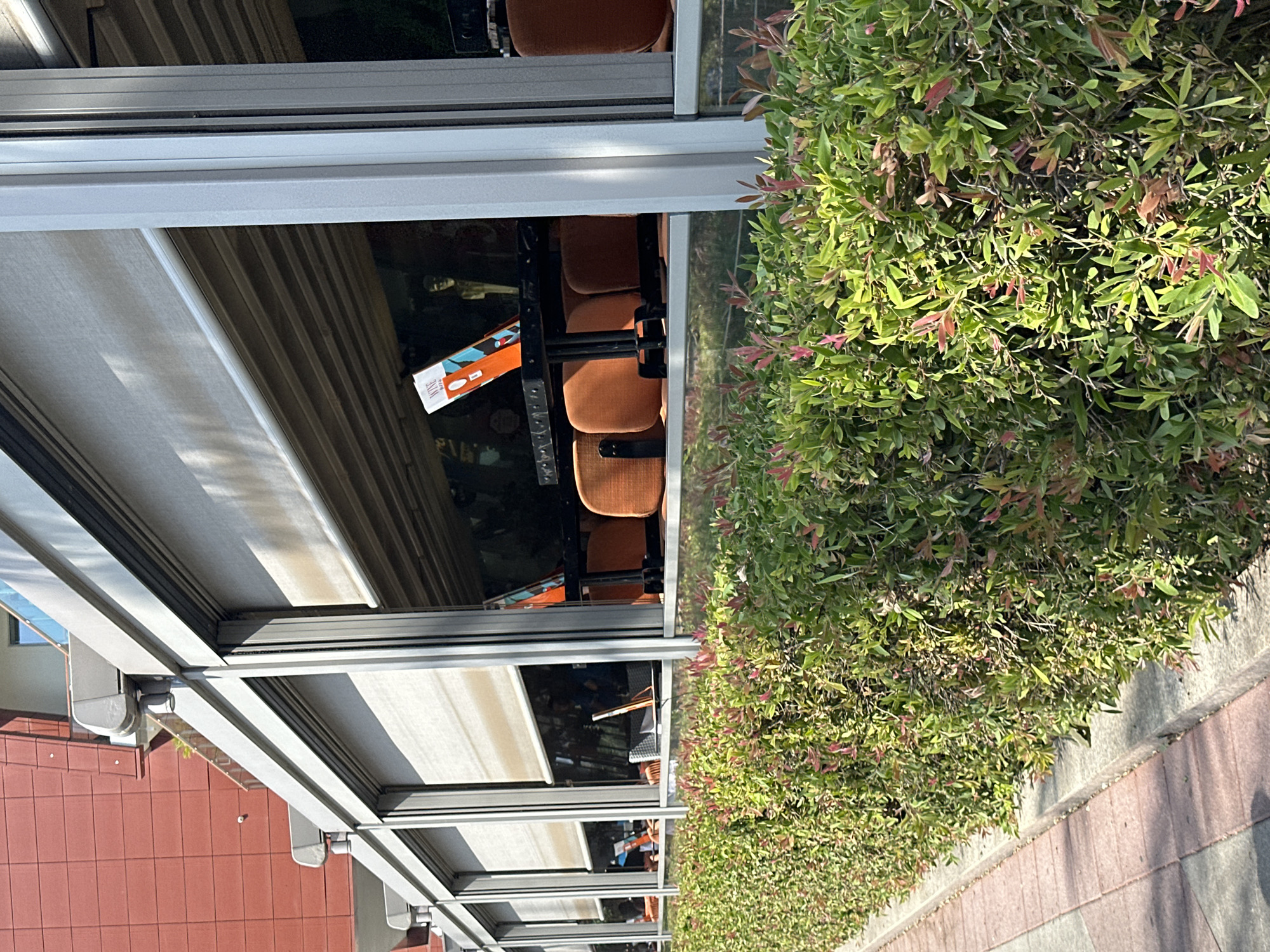 This image depicts the exterior of a cafe or restaurant located in Budva, Montenegro. The establishment features a covered outdoor seating area with retractable blinds that are partially lowered. Inside, tables and chairs, many of which are orange upholstered, are visible. One table has a menu or pamphlet standing upright on it, displaying the word "WINE". Behind the seating area, a wall with various murals or decorations can be seen, along with what appears to be a statue or mannequin. In the foreground, a well-maintained hedge with green and reddish-pink leaves lines the sidewalk, which is paved with interlocking bricks. A red-tiled building rises in the background, suggesting an urban or semi-urban environment. The lighting indicates it is daytime, likely with sunshine casting shadows.
