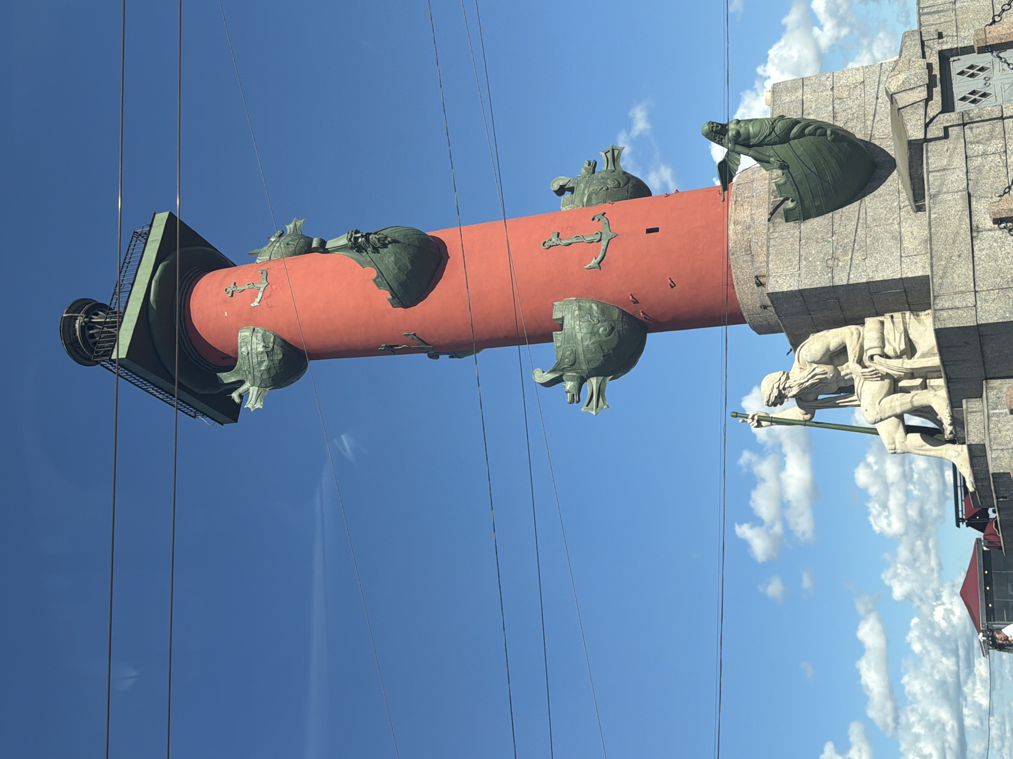 The image depicts the Rostral Columns, iconic landmarks situated on the Spit of Vasilyevsky Island in Saint Petersburg, Russia. The prominent central structure is a tall, bright red cylindrical column topped with a green observation deck. Adorning the column are decorative elements, including bronze ship prows and anchors. At the base of the column, there is a stone plinth. To the left of the column, a large, light-colored sculpture of a seated male figure holding a staff is visible. To the right of the column, a bronze sculpture of a winged mermaid in a ship prow is mounted on the plinth. The sky is a clear blue with scattered white clouds. Several dark, thin wires run horizontally across the frame, suggesting the presence of overhead power lines. In the lower left portion of the image, a red-roofed structure and a glimpse of a person in a white shirt are visible, hinting at the presence of a nearby building or event.