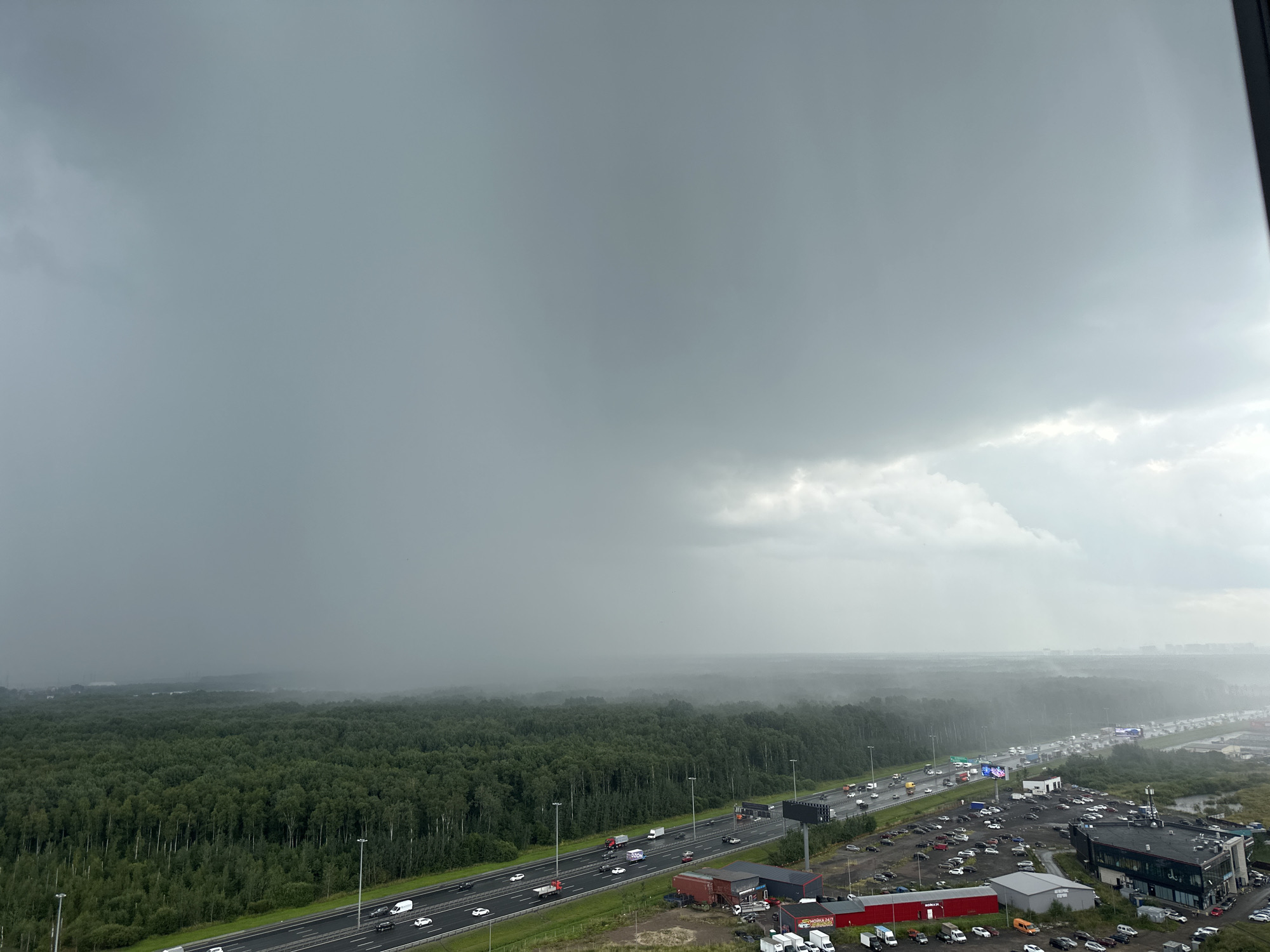 An elevated, wide shot captures a metropolitan landscape under a heavy, overcast sky. The foreground features a multi-lane highway populated by a moderate flow of vehicles, including cars, trucks, and vans. Adjacent to the highway, a large parking lot is filled with numerous cars. Beyond the highway lies a dense, green forest that stretches towards the horizon. In the distance, a hazy layer of mist or fog partially obscures a cityscape, suggesting it is either a developed area or industrial zones. The sky is predominantly a uniform gray, with some lighter patches suggesting diffused sunlight or thinning clouds. The overall atmosphere is muted, indicative of impending or ongoing rain. The scene appears to be taken from a tall building or structure, looking out over the urban fringe and natural landscape. The location is identified as Imeni Sverdlova, Russia.