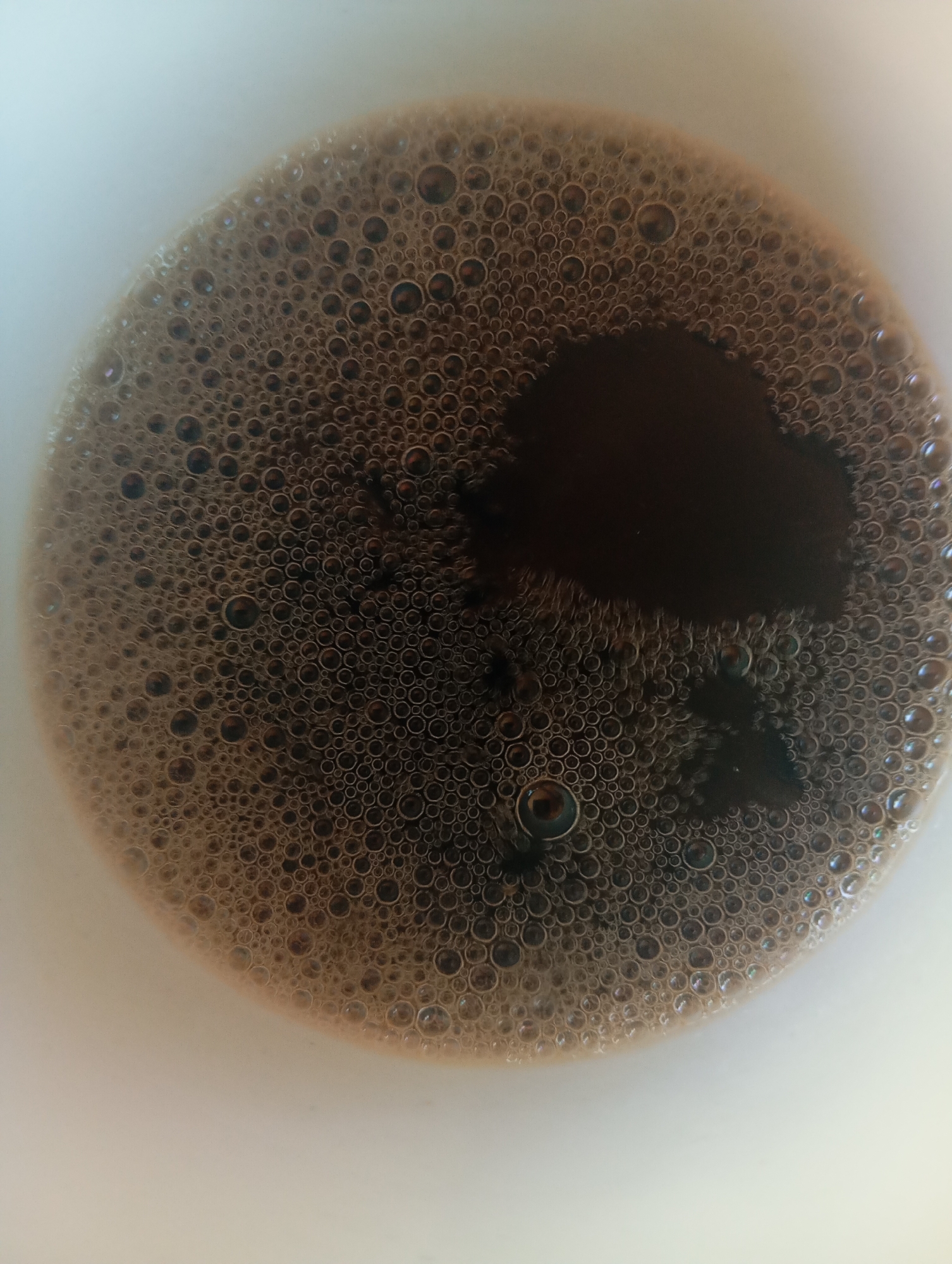 This is a close-up, top-down view of the surface of coffee in a white mug. The coffee has a thick layer of small, dark brown bubbles covering most of the surface. There is a darker, more liquid area in the upper right quadrant, suggesting that the coffee is not uniformly mixed. The mug appears to be made of white ceramic. No specific location in Orenburg, Russia, can be determined from this image.