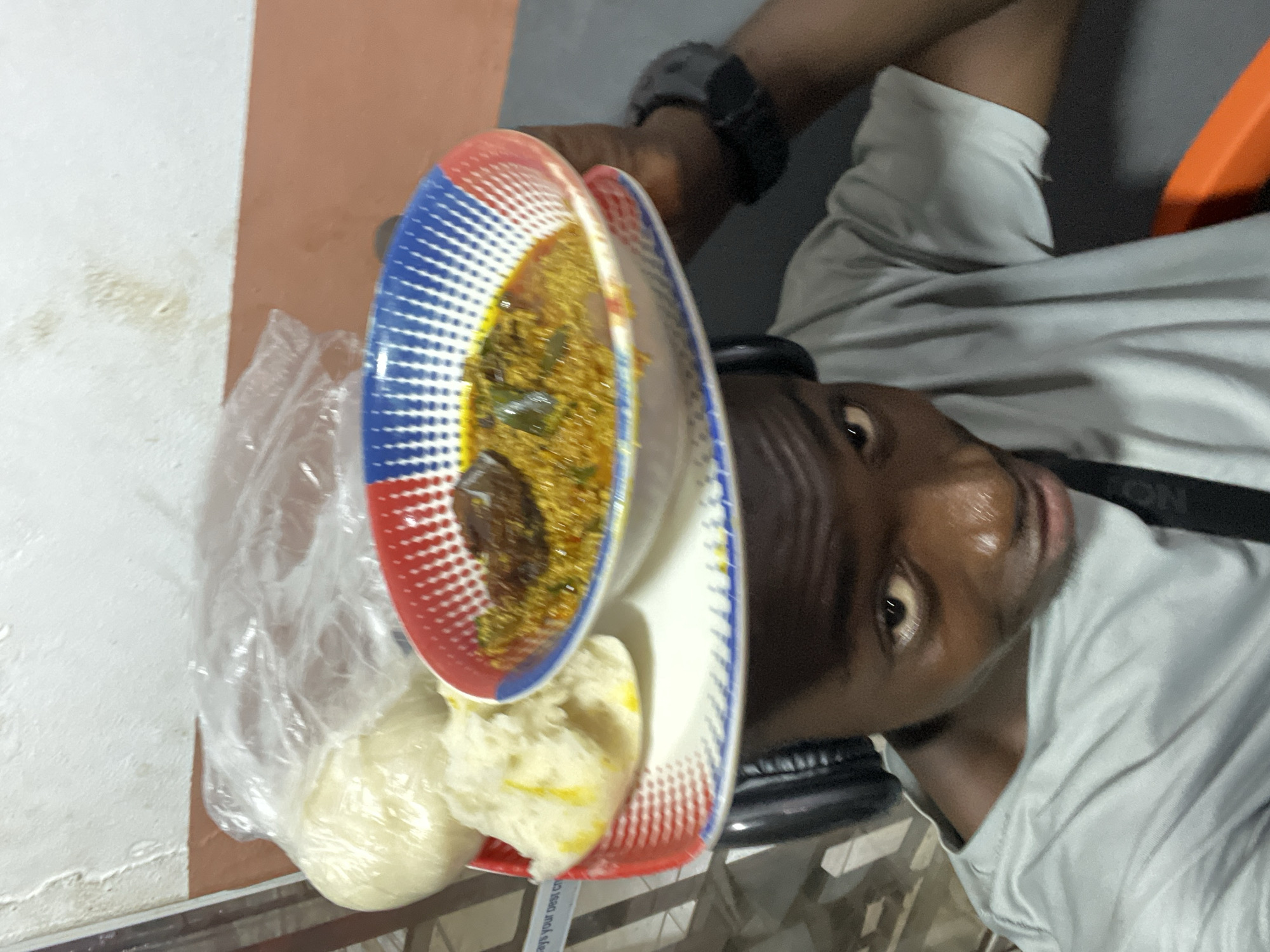 The image, rotated 90 degrees clockwise, captures a male presenting a meal. The person, with dark skin, is visible from the chest up on the right side of the frame, looking directly towards the viewer with a neutral, engaged expression. He wears a light-colored short-sleeved shirt and a dark watch on his left wrist.
In the center, the person holds two stacked plates. The top plate, featuring a distinctive red, white, and blue dotted pattern, contains a reddish-brown stew or soup, which appears to have leafy greens and possibly pieces of protein. Below this, on the lower plate, is a clear plastic bag holding a serving of white, dough-like food, likely a swallow staple such as fufu or pounded yam, common in Nigerian cuisine.
The background consists of a simple wall, segmented into an upper white or off-white section and a lower reddish-brown or orange band. A table or counter edge is partially visible. The lighting is ambient, suggesting an indoor setting. Faint, largely illegible text can be seen on a dark strap or surface near the bottom edge of the image. The scene depicts a common way a meal might be served or presented in the given context of Nkpor, Nigeria.