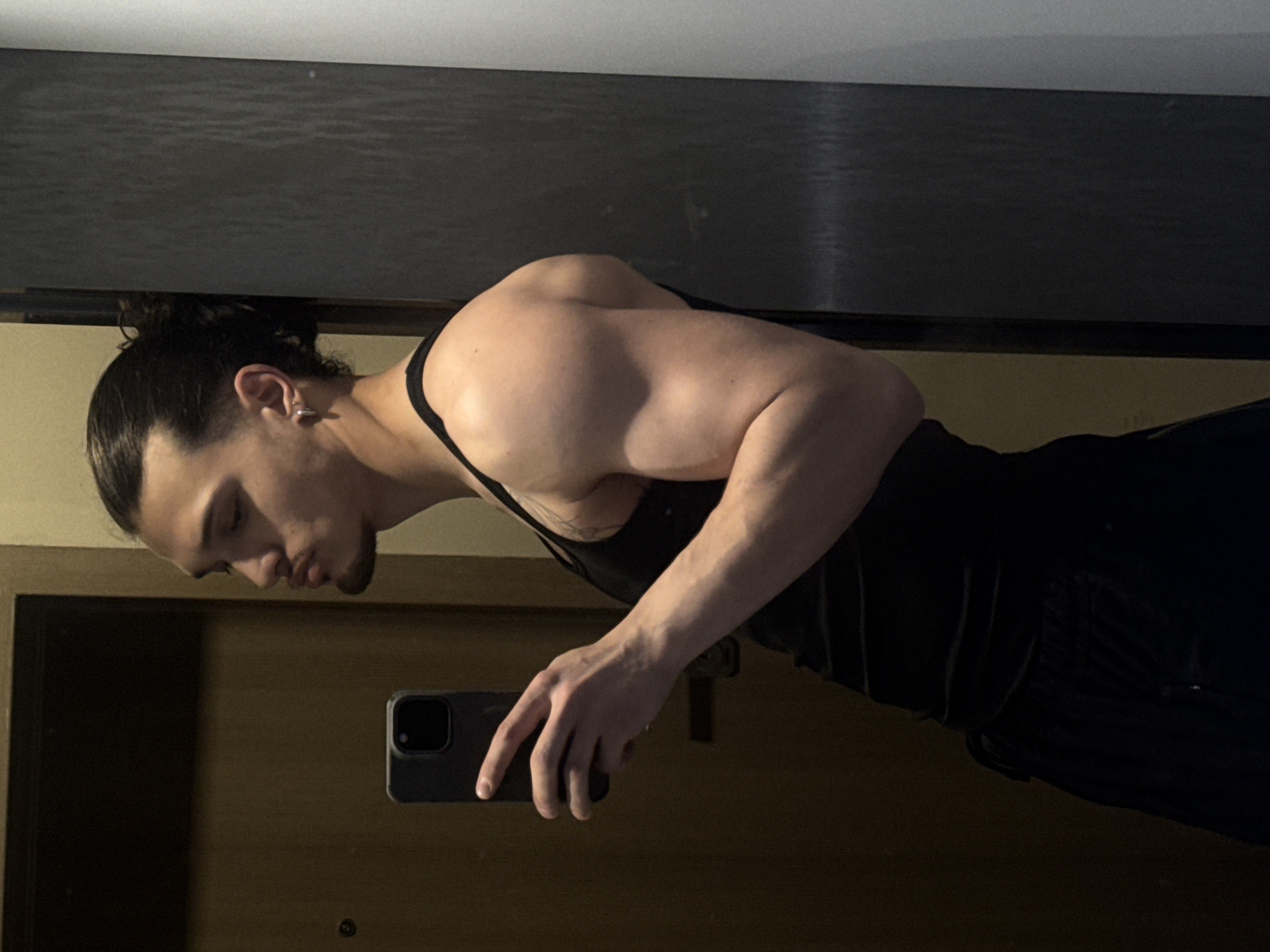 This image, captured in Sofia, Bulgaria, features a young, muscular man taking a mirror selfie. The photo is rotated 90 degrees counter-clockwise. The man has dark hair pulled back into a bun or ponytail and a light beard. He wears a black tank top and dark pants/shorts, showcasing his well-defined physique, including prominent biceps, triceps, and shoulders, with visible veins on his forearms. He has a silver hoop earring in his left ear.
He holds a dark-colored smartphone, likely an iPhone, in his right hand, pointing it towards himself. His head is tilted downwards, and his eyes appear closed or looking down, conveying a pensive or thoughtful expression.
The background suggests an indoor setting, possibly a room. Above the man (if correctly oriented) is a dark, textured surface, while below him is a lighter, possibly wooden or paneled surface. The lighting is dim, indicating evening or night, or an interior space with low light levels. There is no visible text in the image.