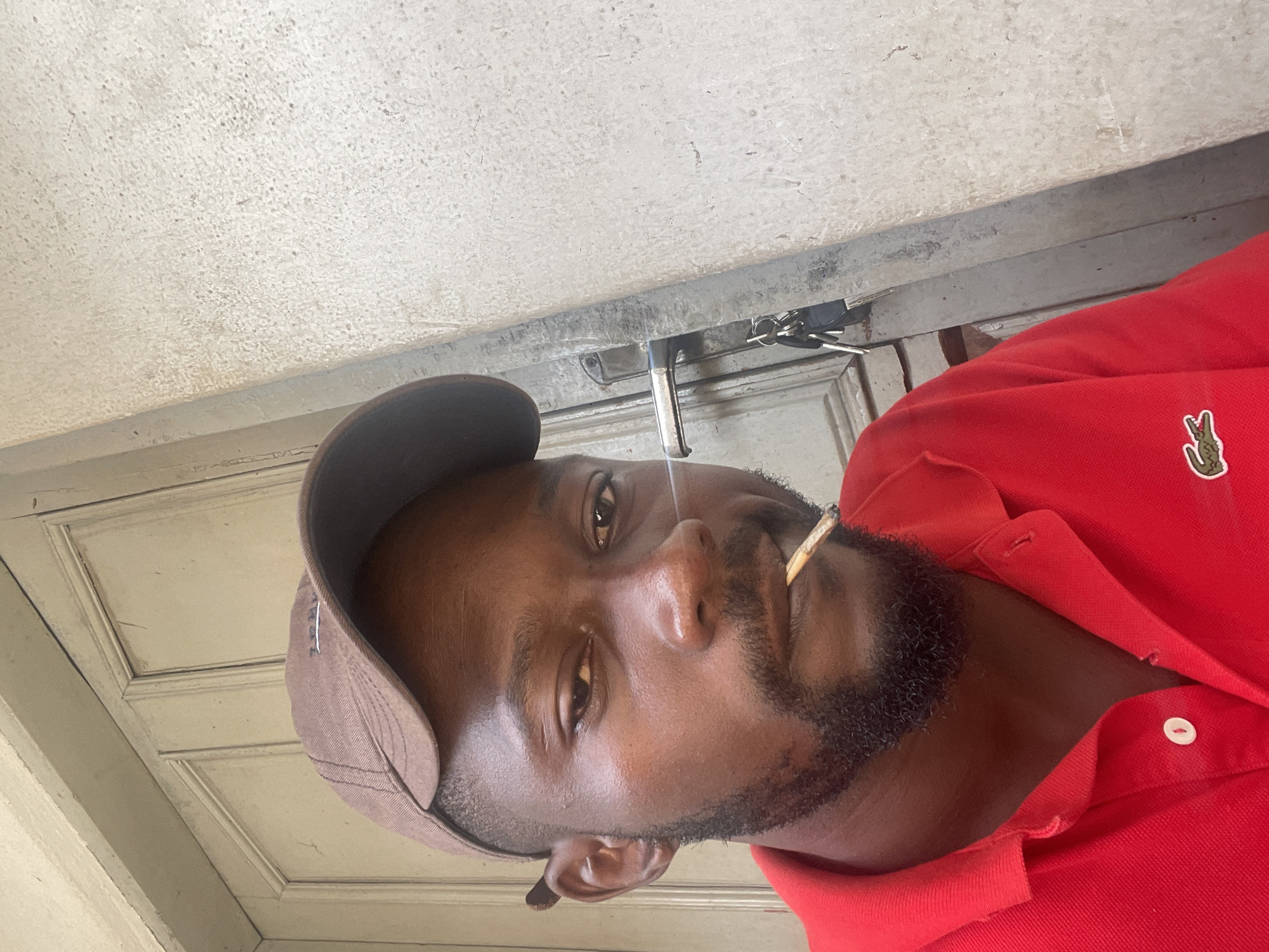 A man wearing a red polo shirt and a brown baseball cap is seen from the chest up, smoking. He has a beard and is looking towards the camera. Behind him is a light-colored door and a textured wall with a door handle and keys. The scene appears to be outdoors, likely in front of a building.