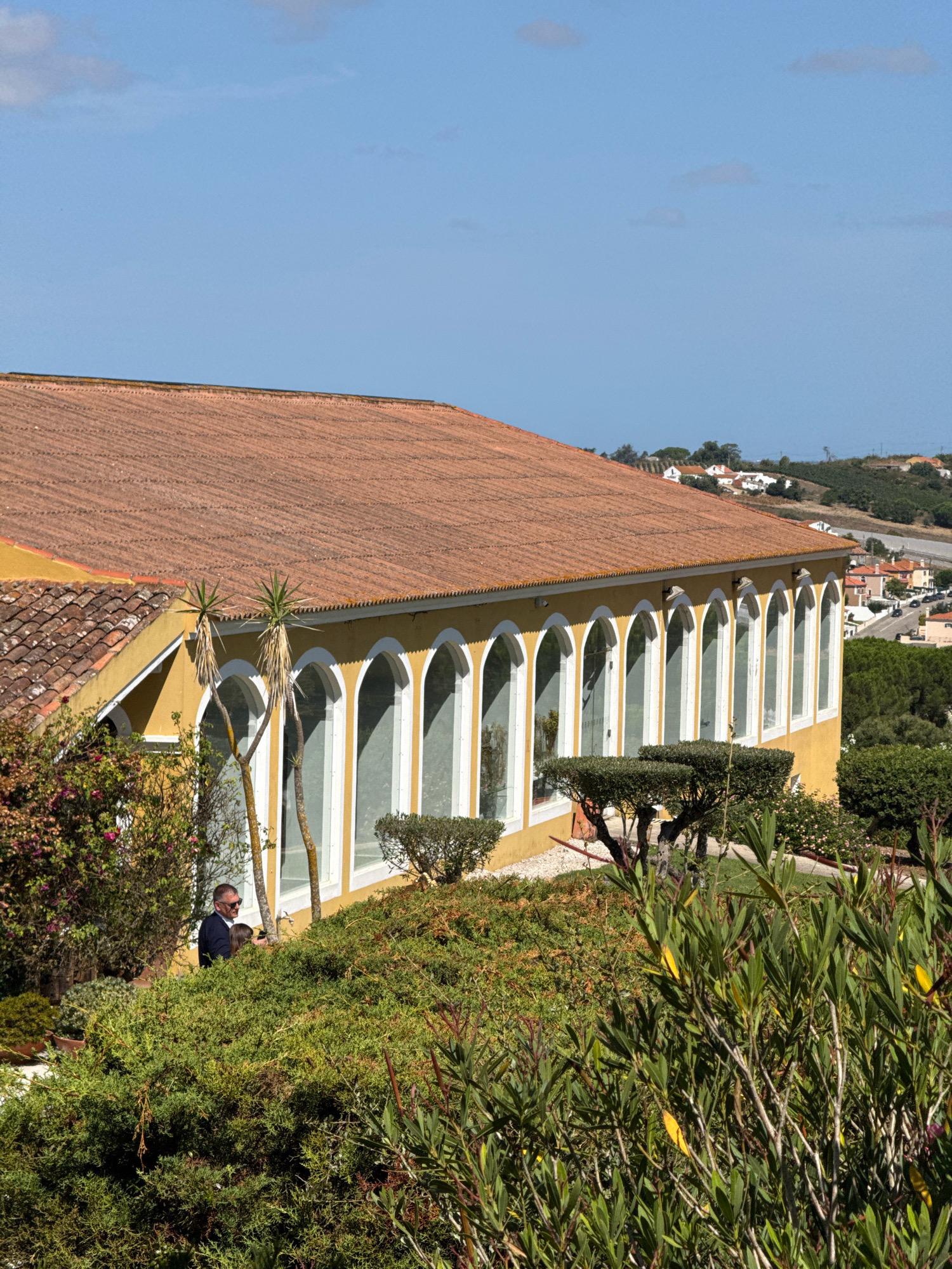 The image shows a long, yellow building with a terracotta tiled roof, featuring a series of arched windows. The building appears to be situated on a hillside with lush green vegetation in the foreground. In the middle ground, a man wearing sunglasses and a dark blue shirt is standing near the building, looking away from the camera. Behind the building, a small town with red-tiled roofs is visible, nestled amongst rolling hills. The sky is clear and blue. The architectural style of the building, particularly the arched windows and the terracotta roof tiles, is characteristic of buildings found in the region of Mafra, Portugal, known for its historical palaces and traditional Portuguese architecture.